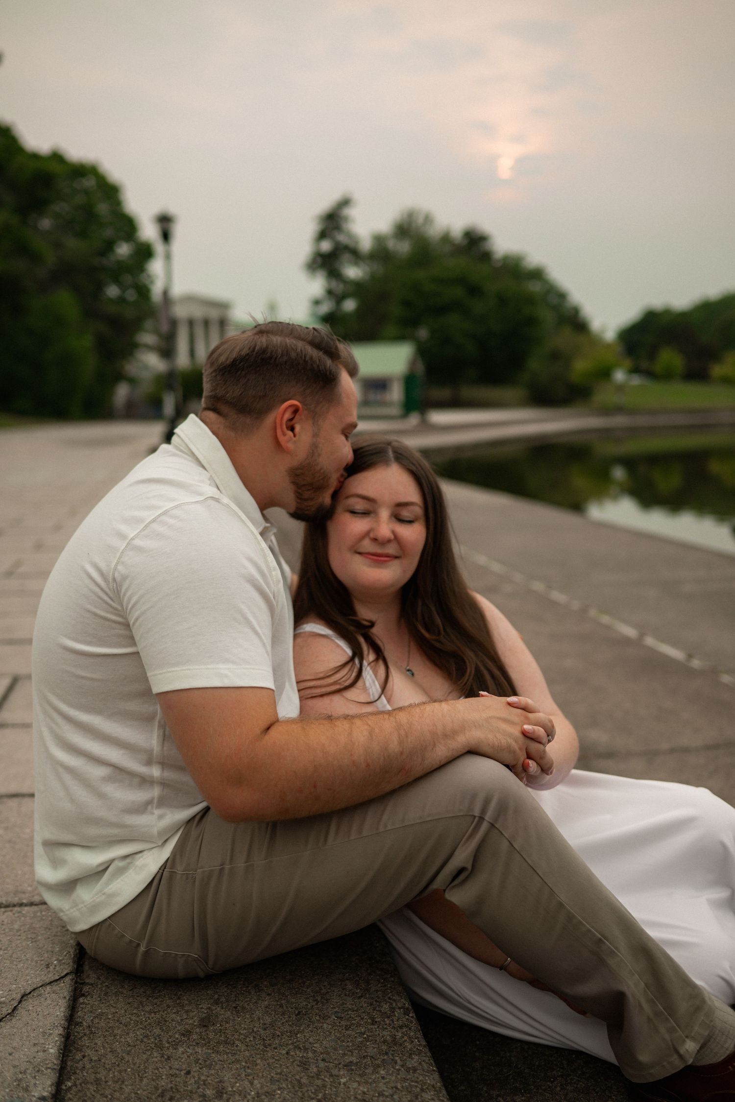 Couple sitting together on stone path by a pond at dusk, man kissing woman's temple as she smiles.