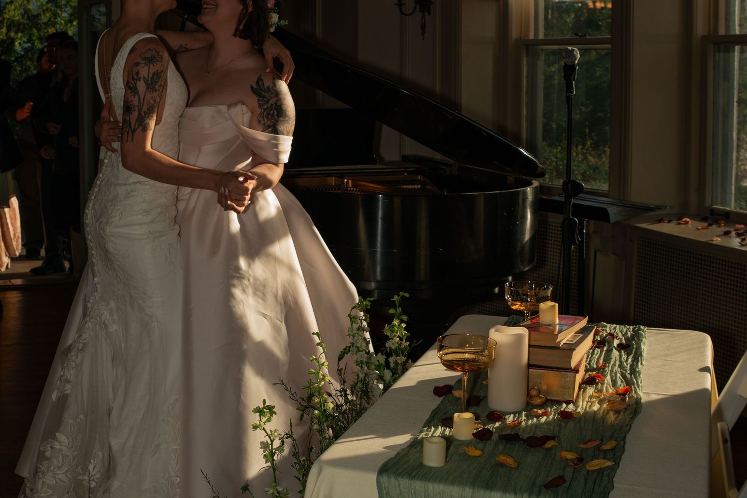Bride in white lace wedding gown standing near elegant cake table with candles and greenery in sunlit venue.