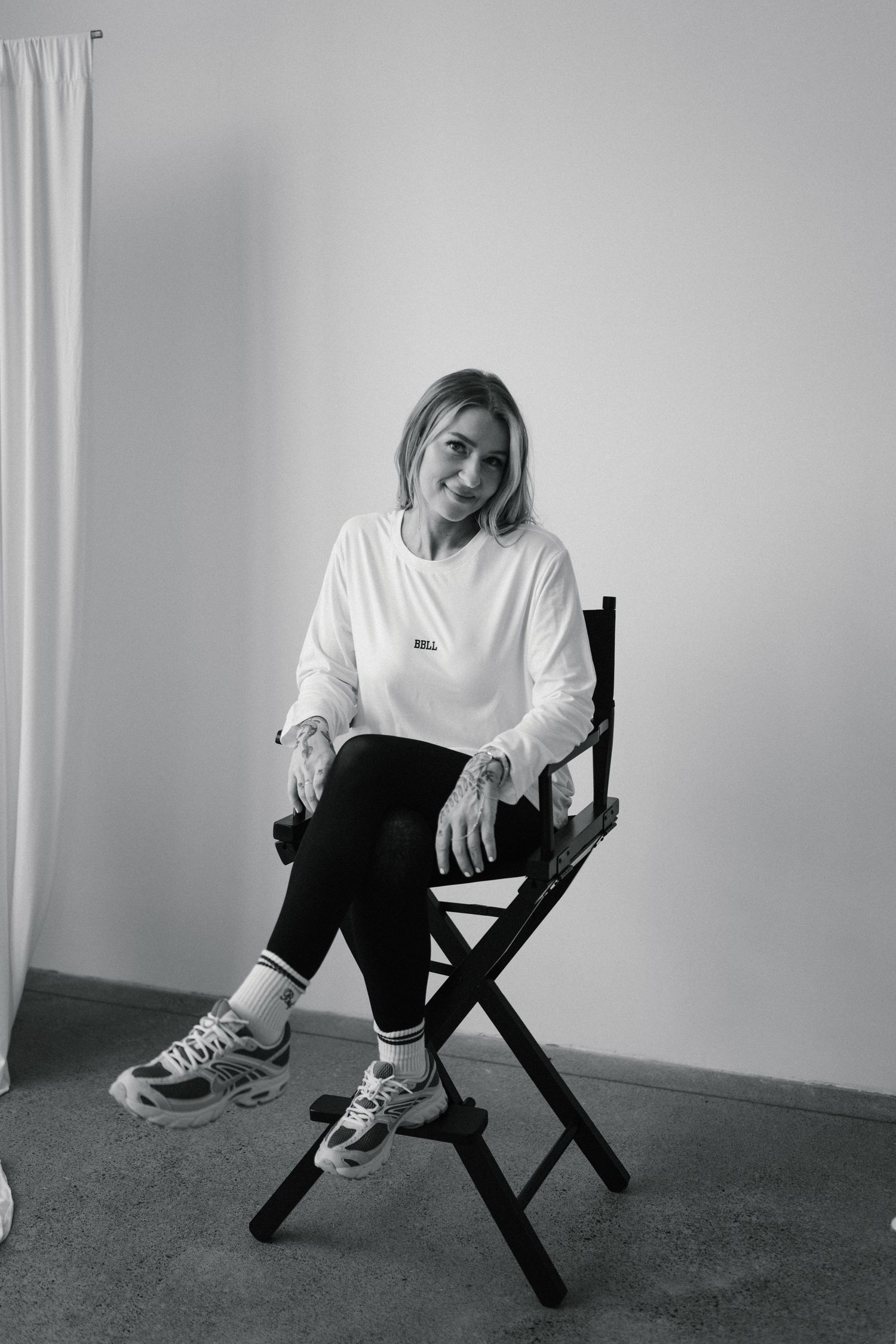A blonde woman sits relaxed in a director's chair wearing a white sweatshirt and black leggings in a black and white photo.
