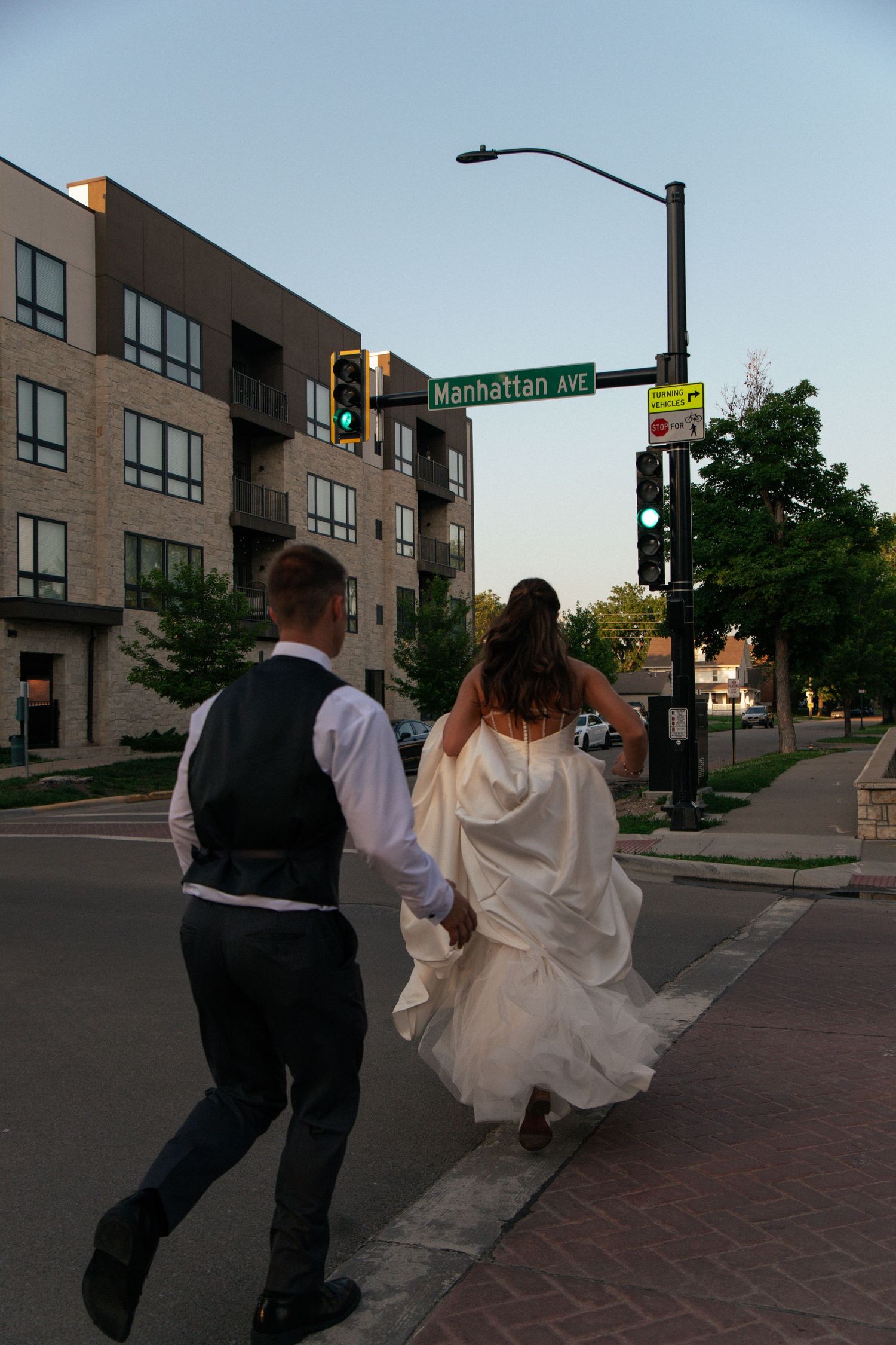 Bride and groom walking hand-in-hand across Manhattan Ave intersection at dusk.