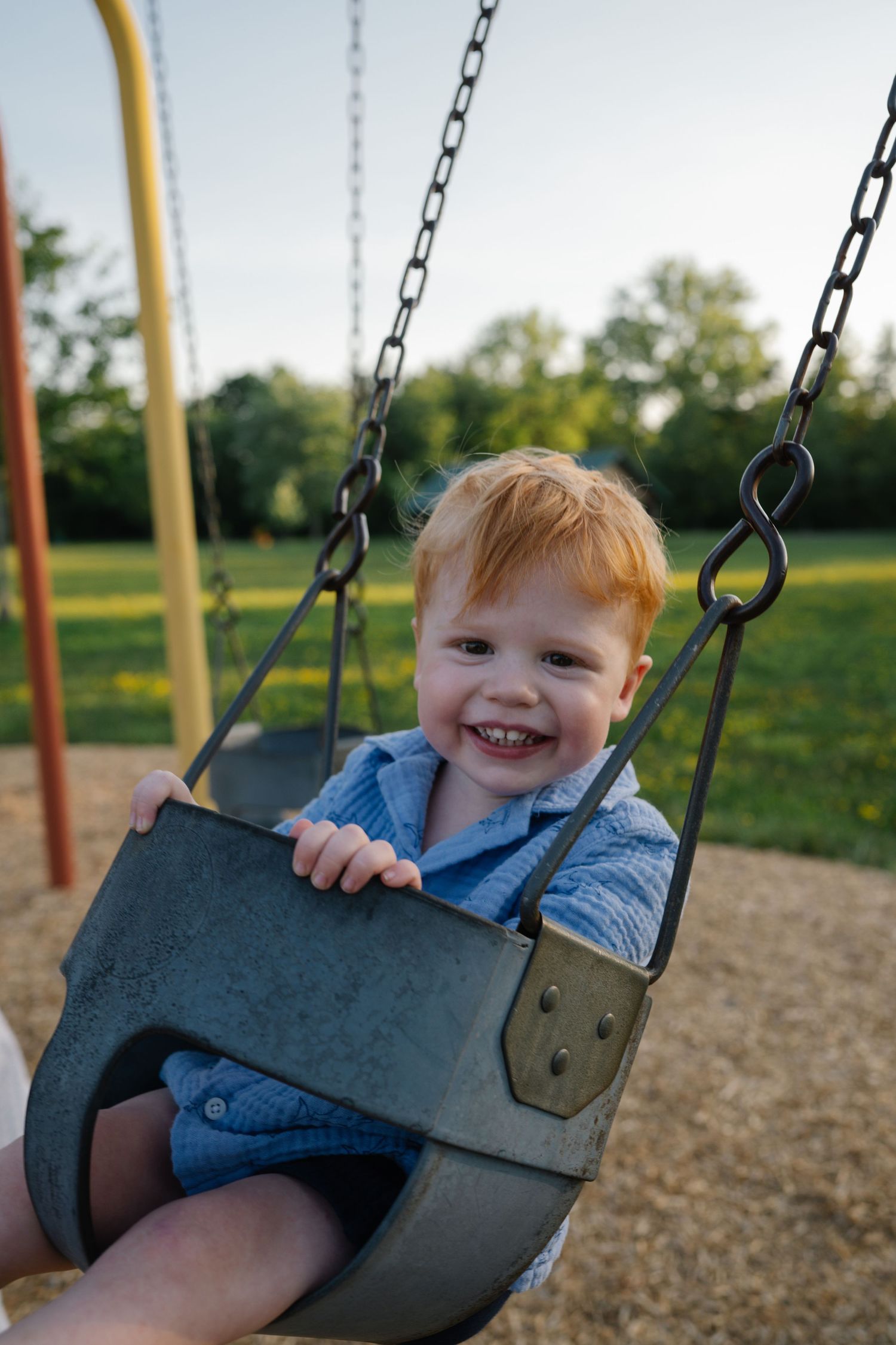 Happy red-haired toddler smiling in a bucket swing at a playground on a sunny day.