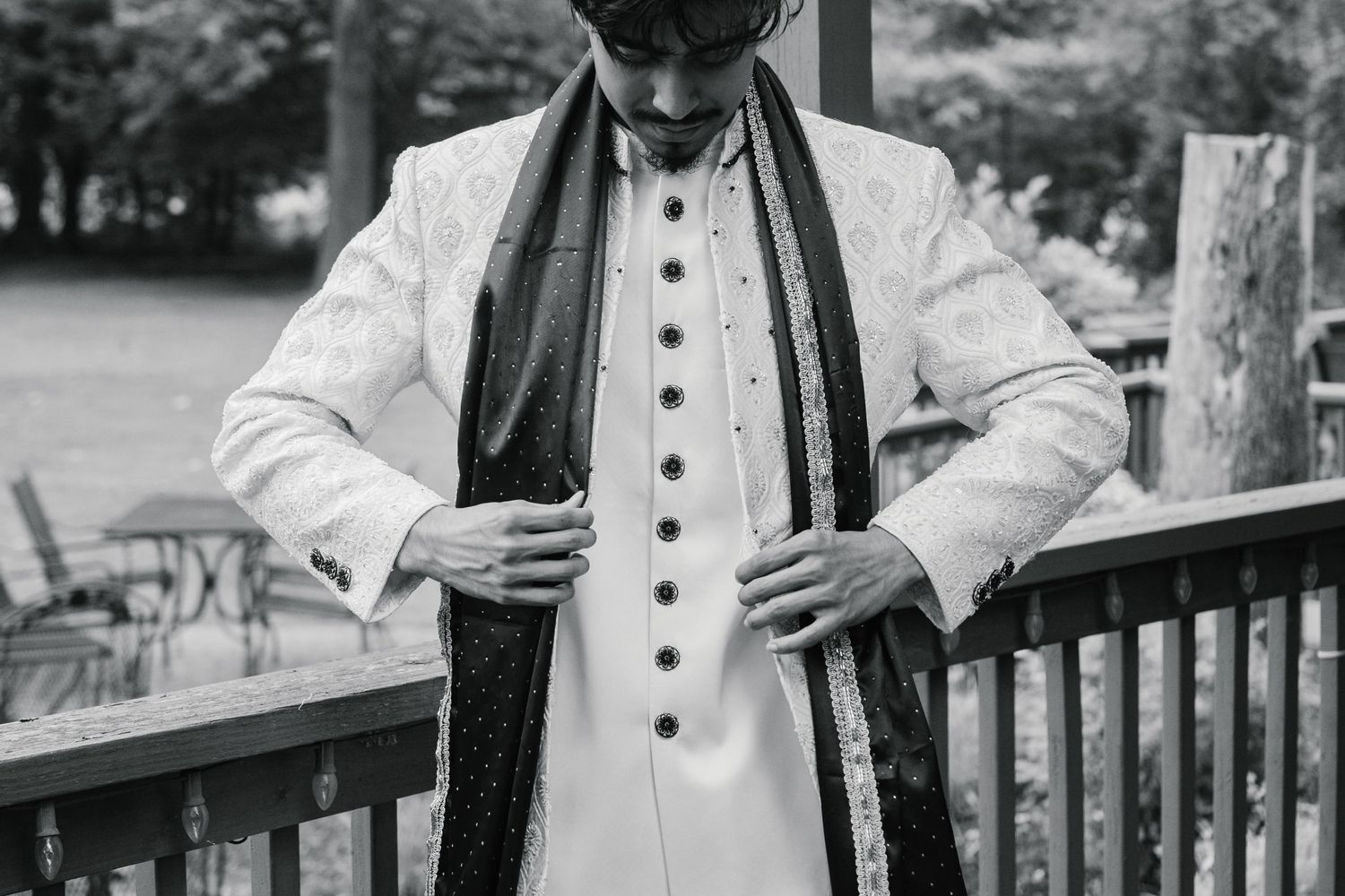 Black and white photo of a man in traditional South Asian sherwani adjusting buttons on a decorated wooden deck.