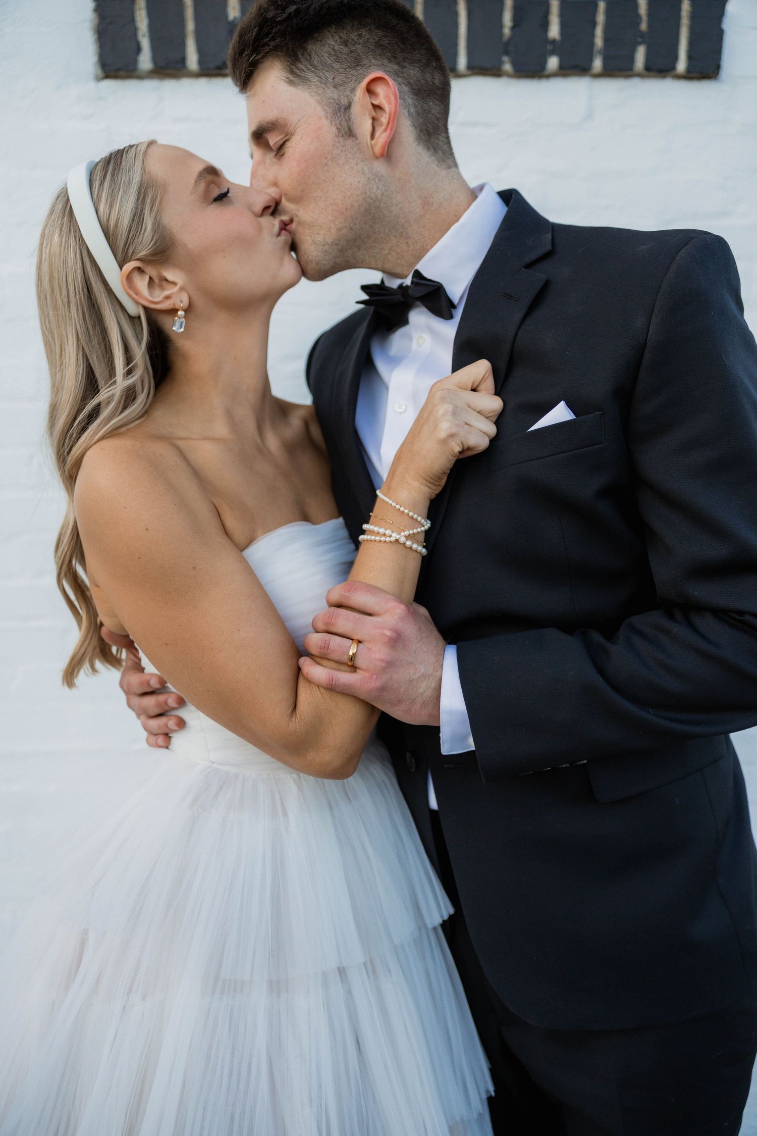A bride in a strapless white gown and groom in a black tuxedo share a romantic kiss against a white brick wall.