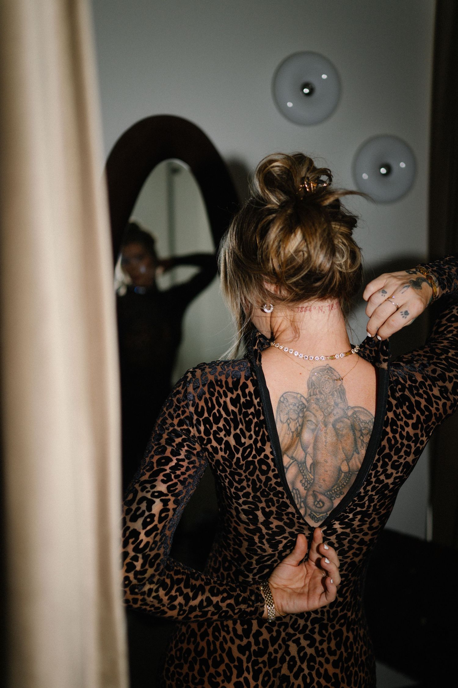 Woman in leopard print backless dress showing elephant back tattoo, adjusting necklace in mirror.