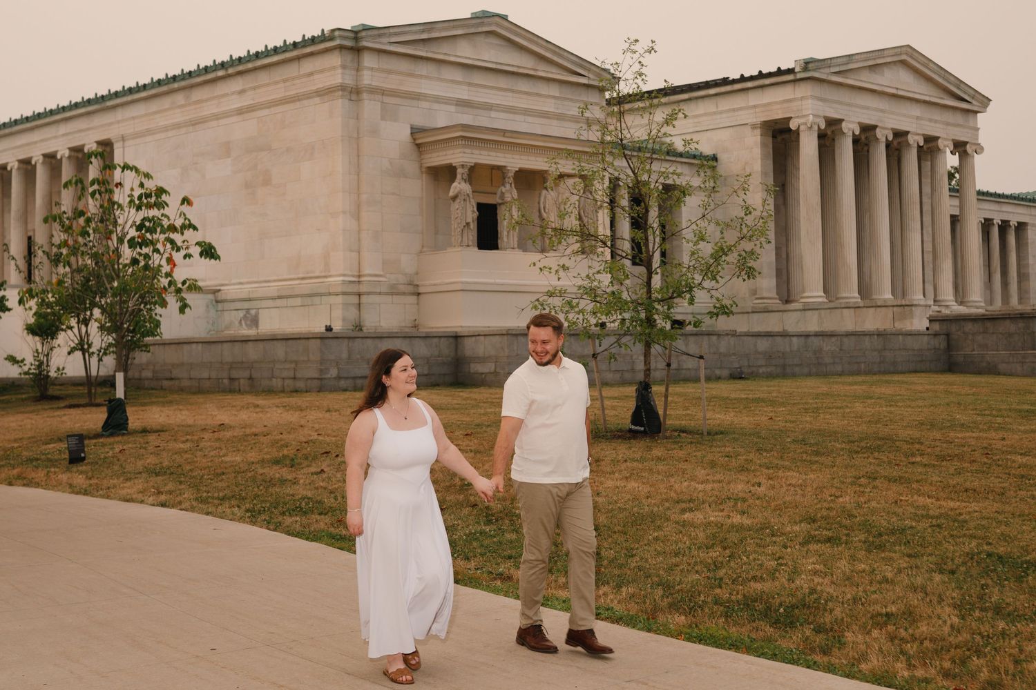 Couple holding hands walking in front of a neoclassical museum building with columns on a hazy day.
