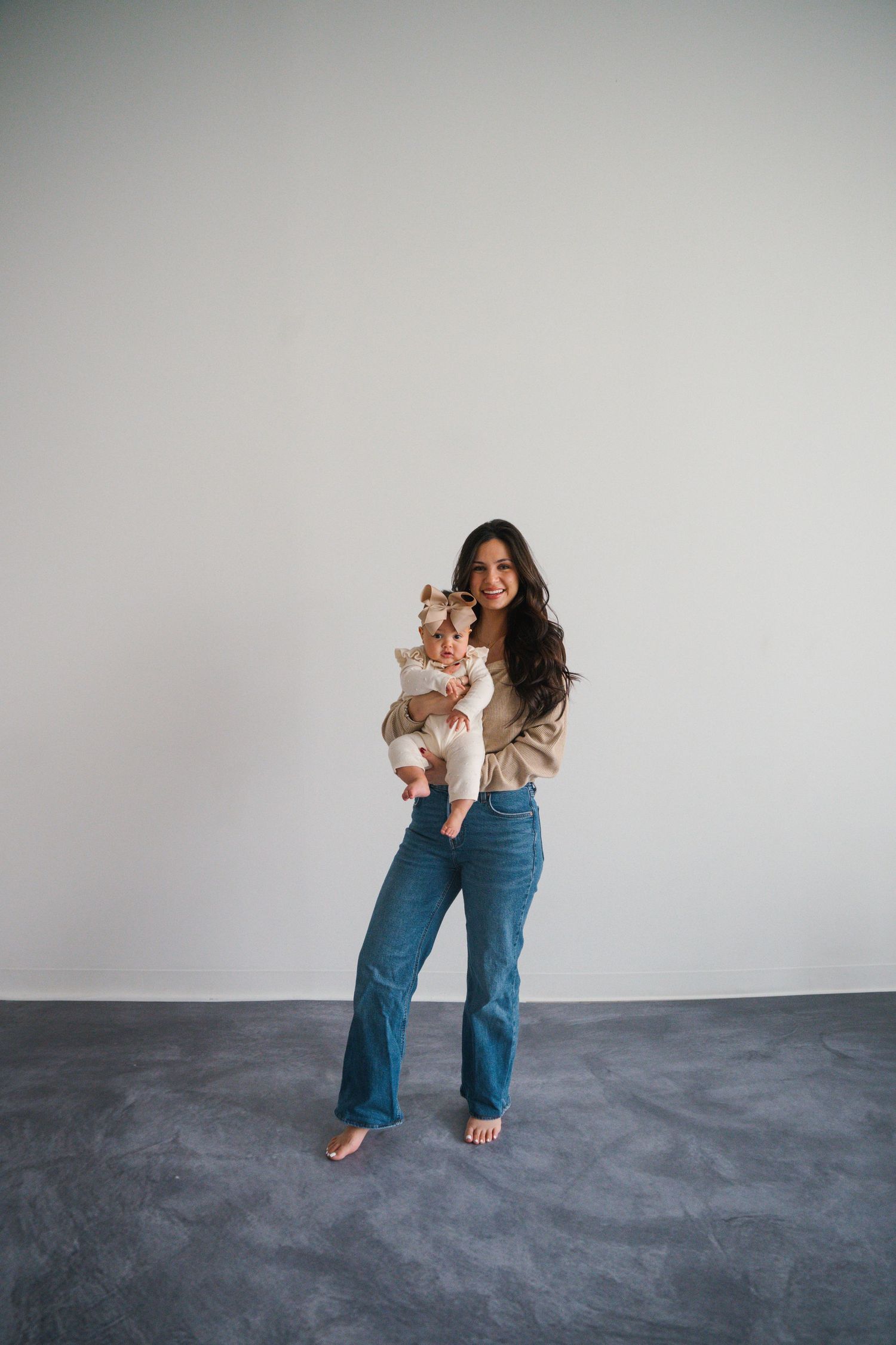 Young woman with long dark hair holding a baby, wearing blue wide-leg jeans and beige top, standing against white wall.