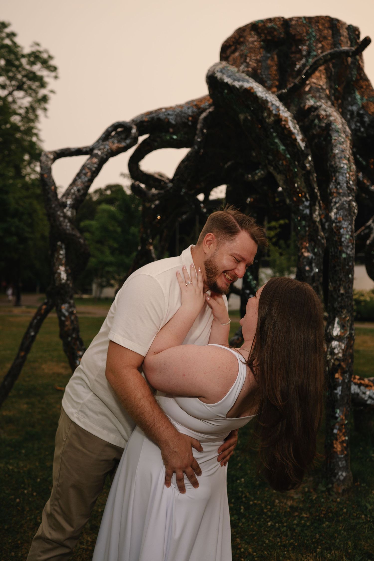 Couple sharing a romantic moment outdoors near a large spider sculpture in a park at dusk.