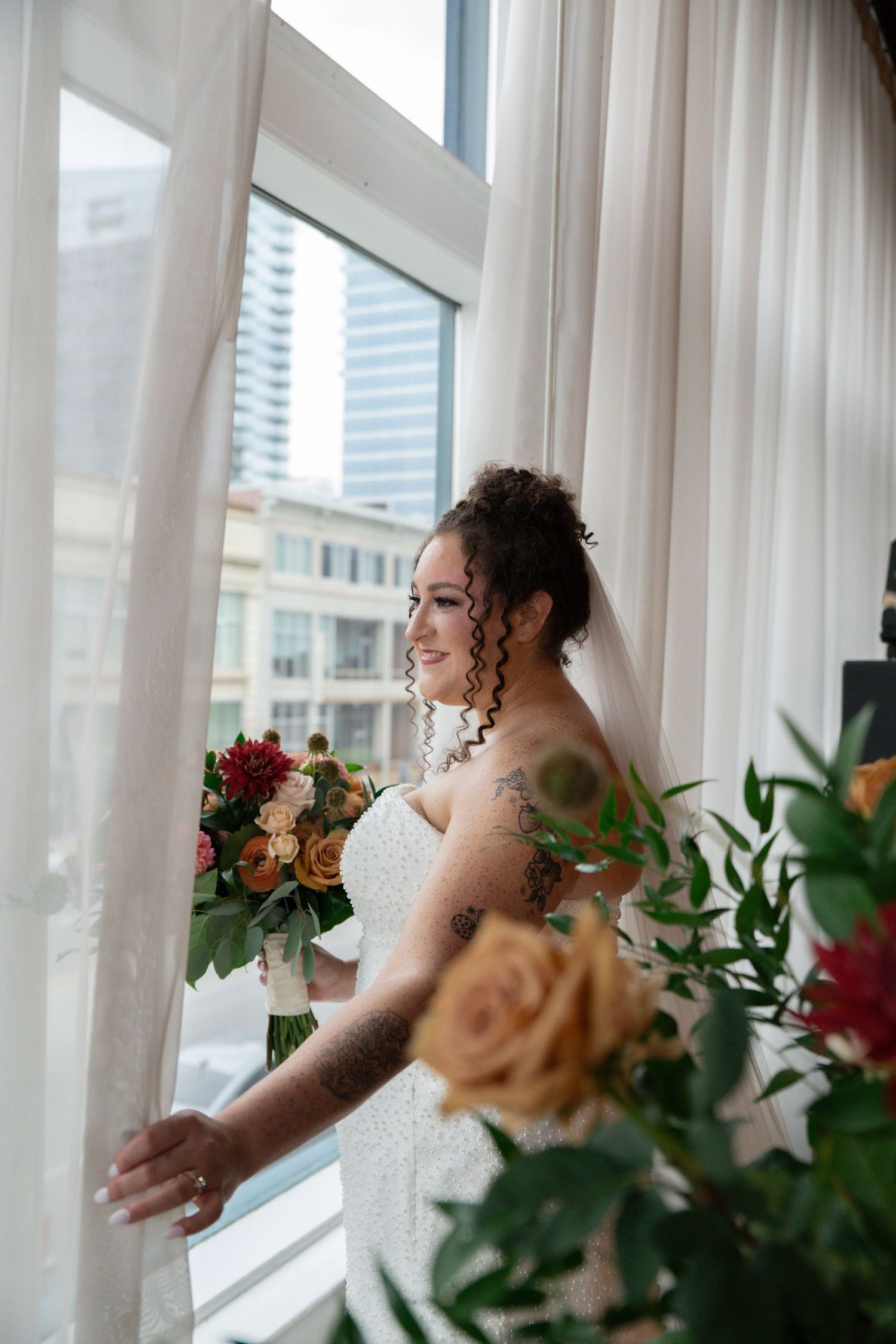 Tattooed bride in white gown holds colorful bouquet by window with city view, wearing veil and updo hairstyle.