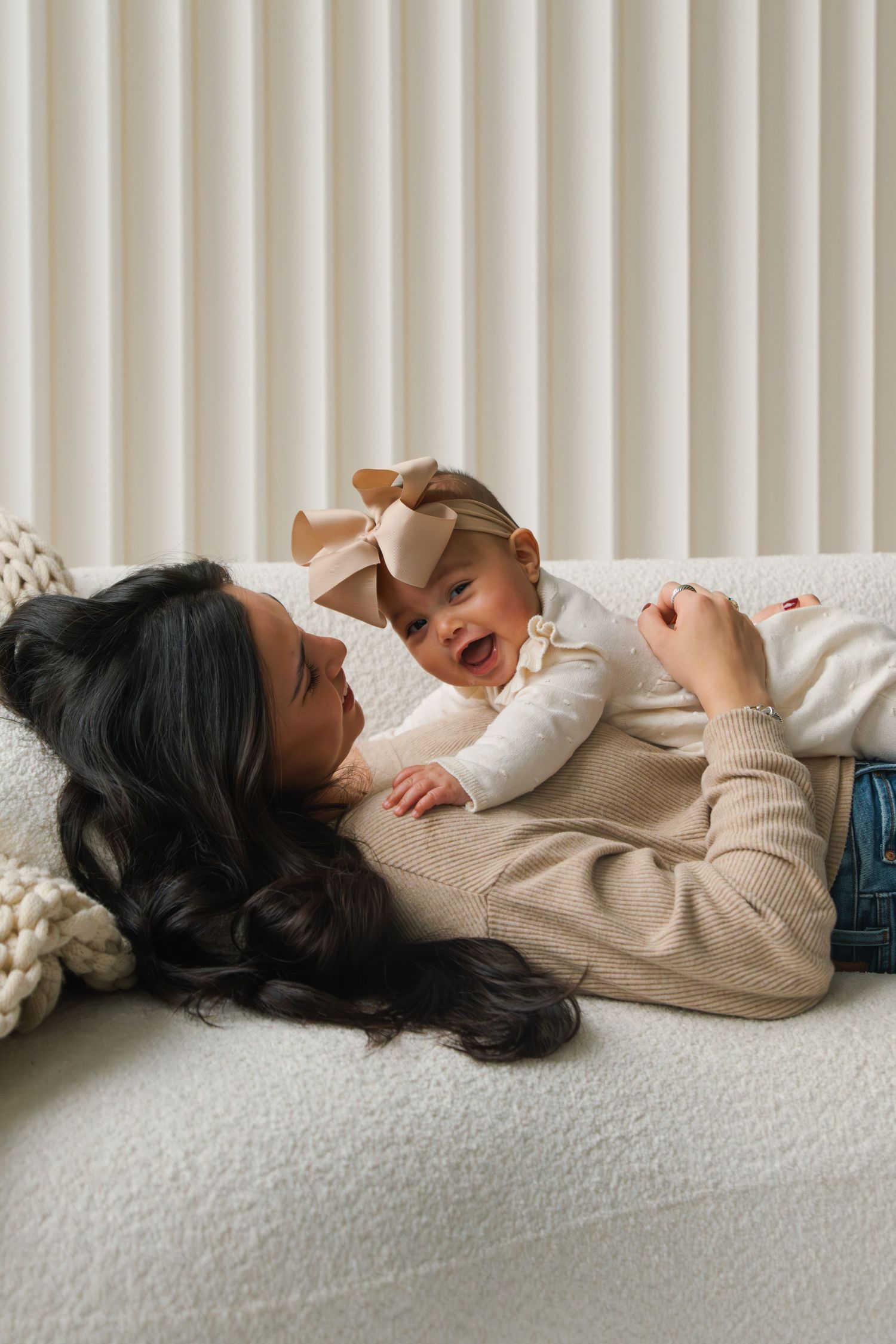 A mother lying on a couch smiling at her laughing baby girl wearing a bow headband.