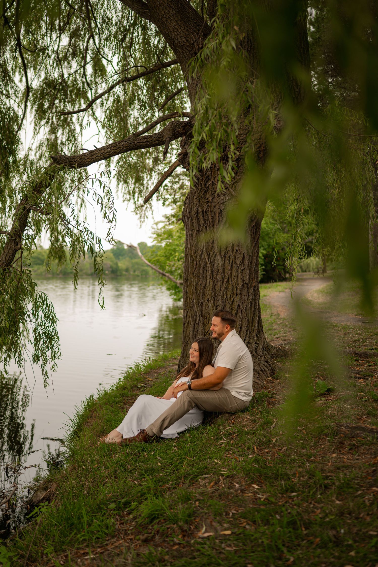 Couple sitting together under a willow tree by a calm river in a lush green park setting.