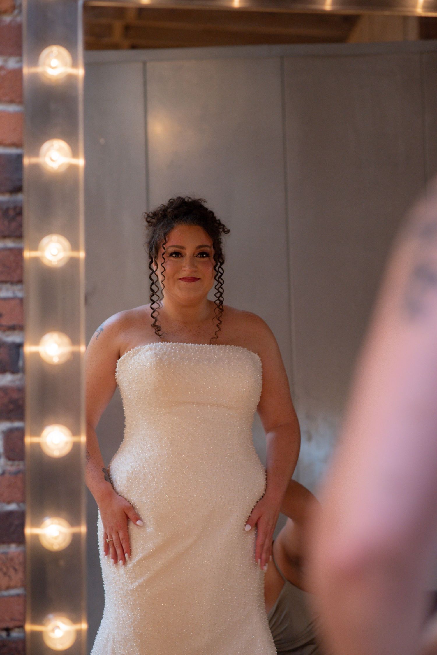 Bride in strapless white beaded gown poses before a Hollywood mirror with glowing bulbs in a brick-walled room.