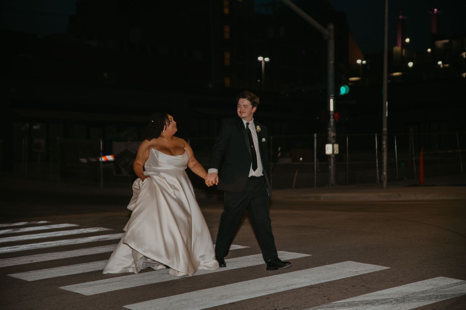 Bride and groom holding hands crossing a zebra crossing at night in a city street, wearing wedding attire.