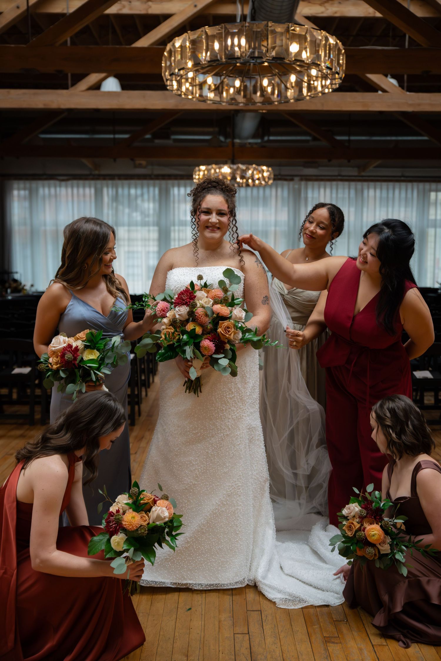 Bride in white lace gown holds floral bouquet surrounded by bridesmaids in burgundy and gray dresses in elegant venue.