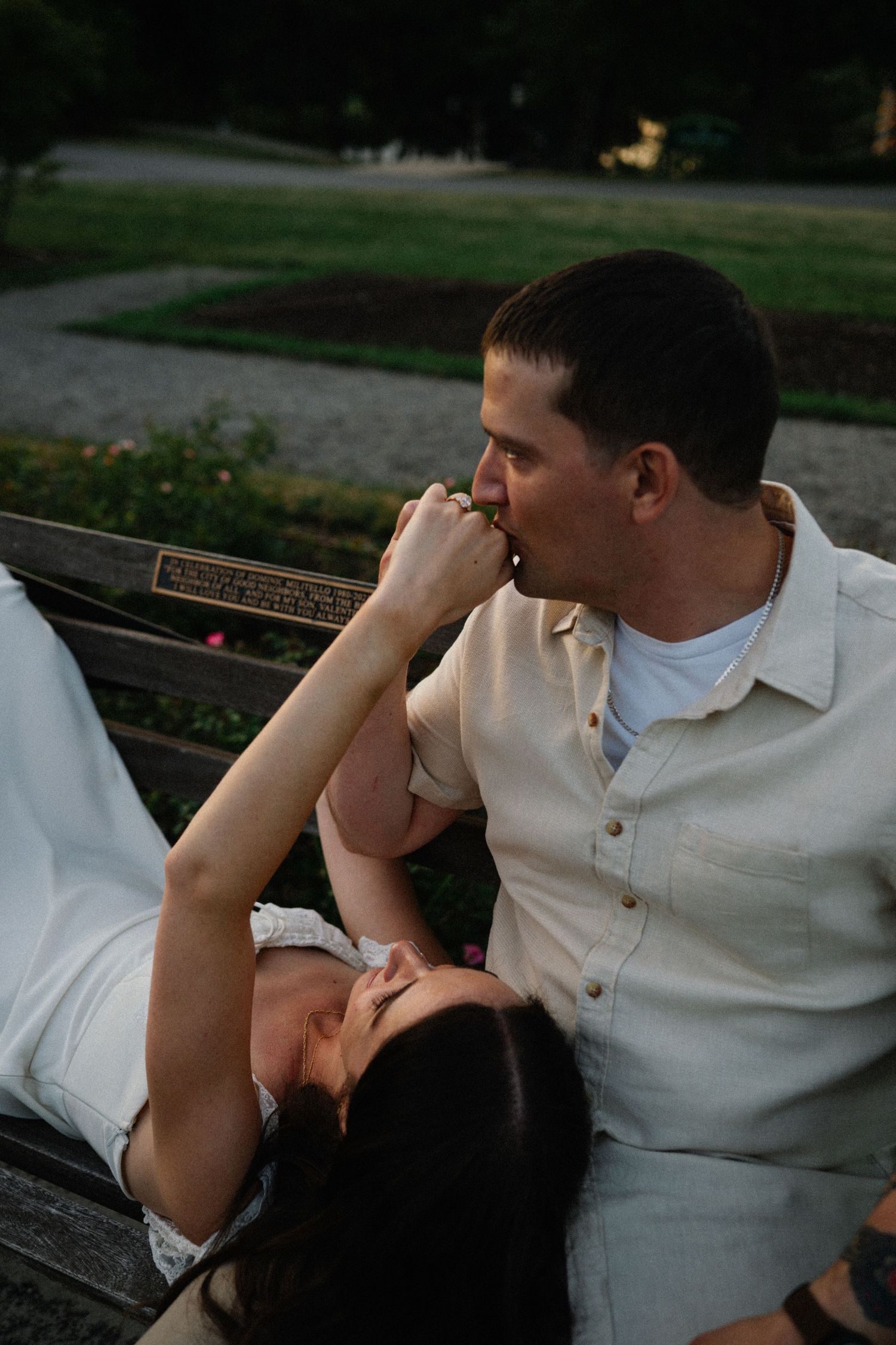 Romantic couple on a park bench, man kissing woman's hand as she reclines, sharing a tender moment outdoors.
