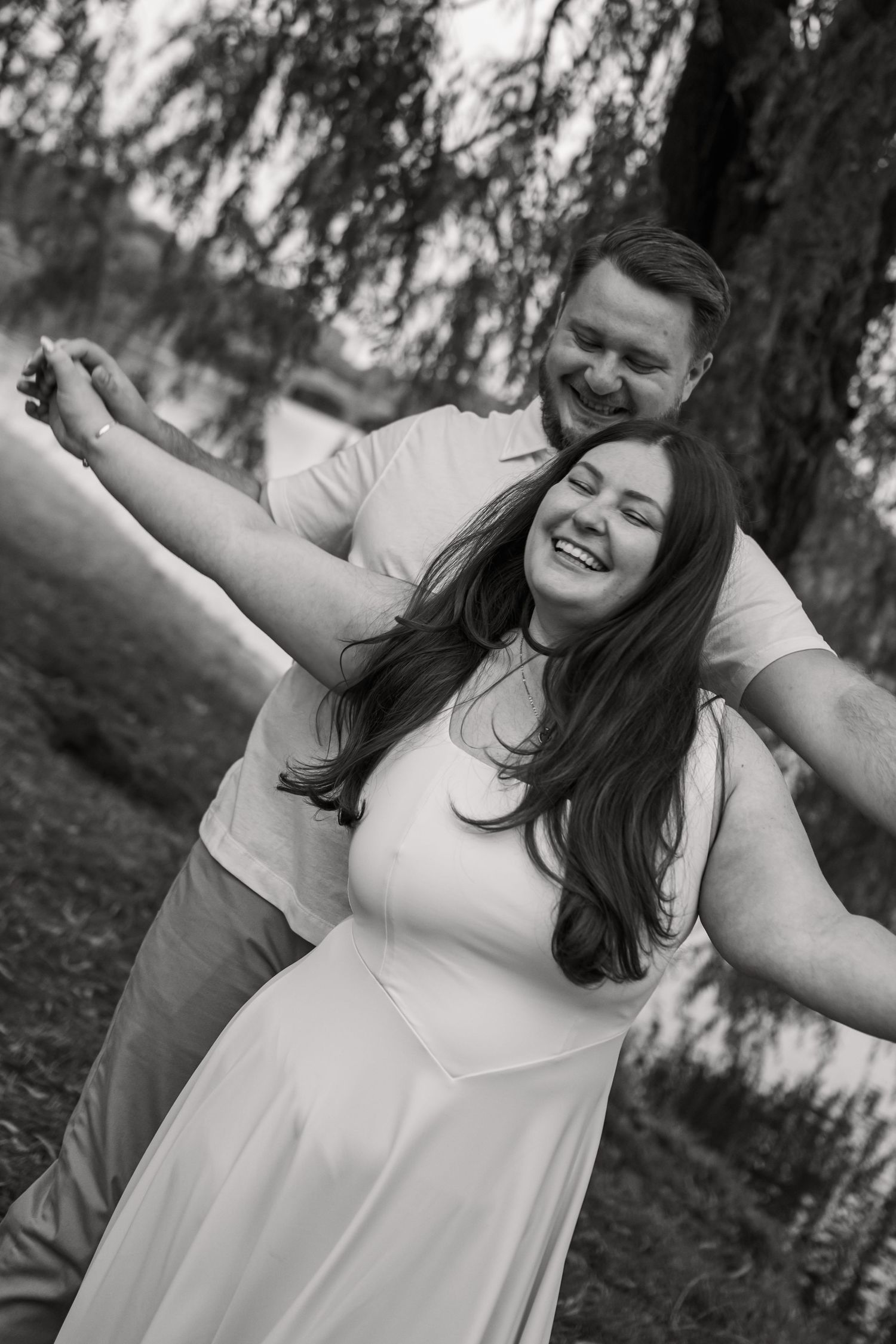 Black and white photo of a joyful couple dancing outdoors near a willow tree, woman in white dress laughing.