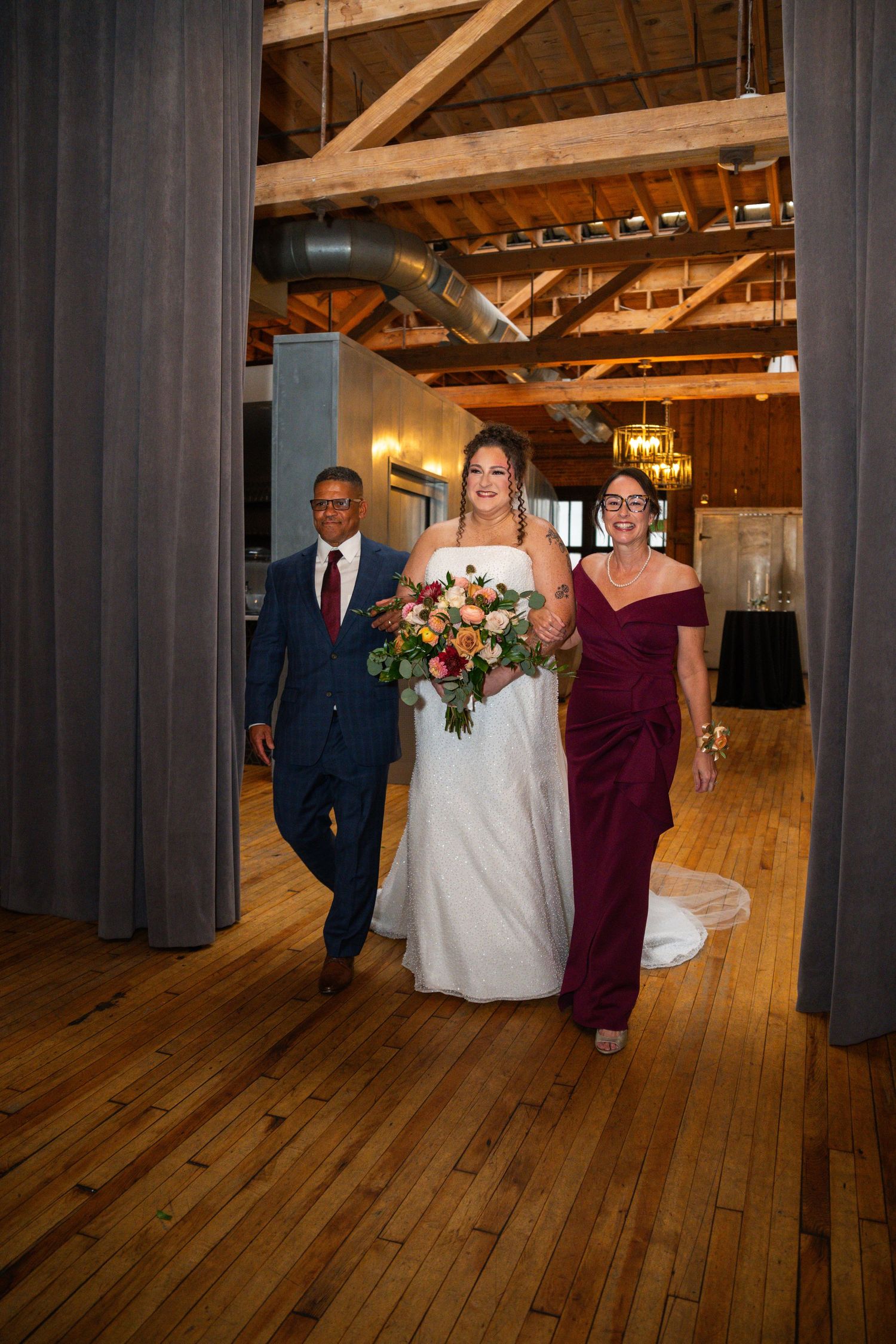 A wedding party walks down a rustic venue hallway with exposed wooden beams and hardwood floors.