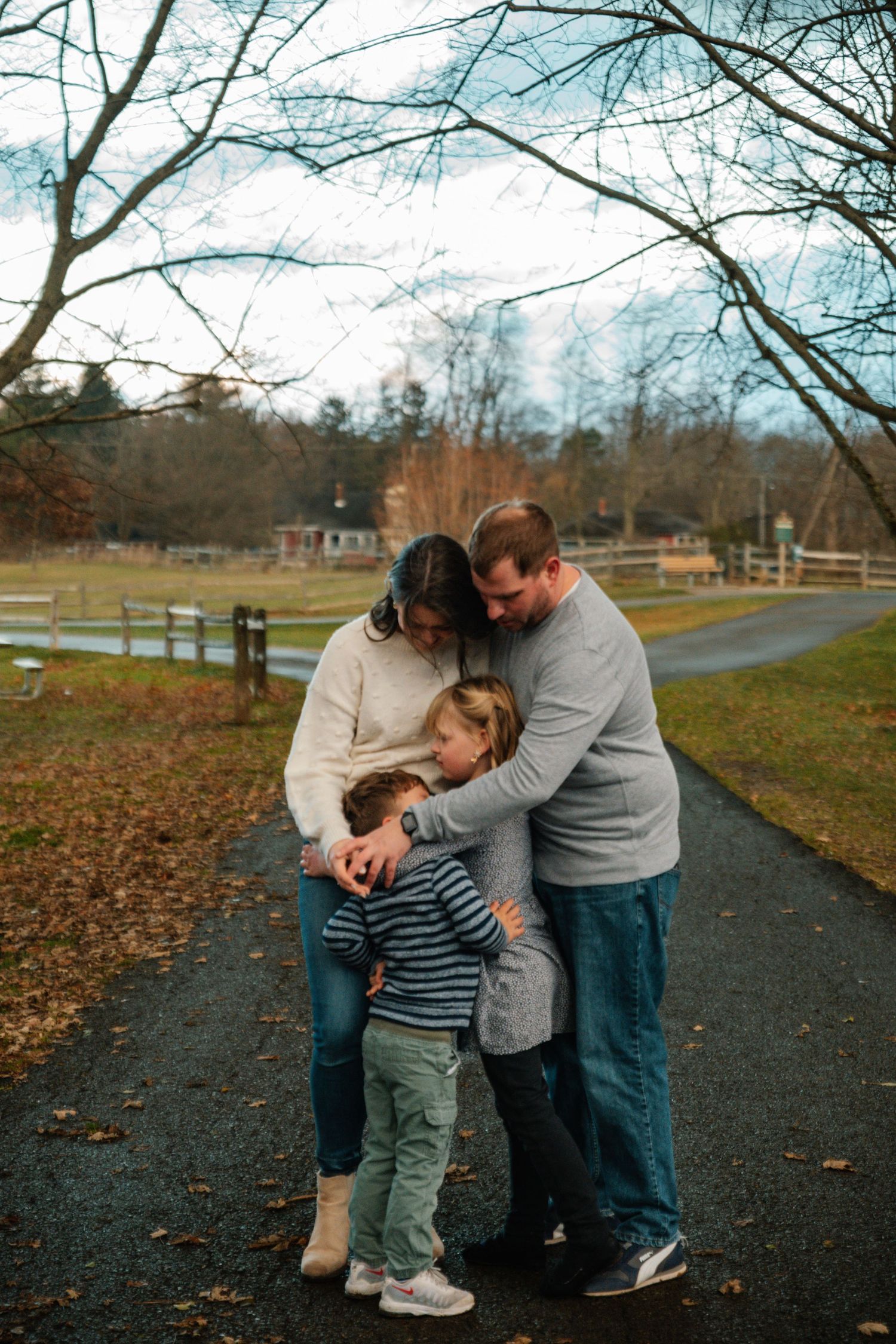 Family of three sharing a warm embrace on a scenic autumn path surrounded by bare trees and a rustic farm.