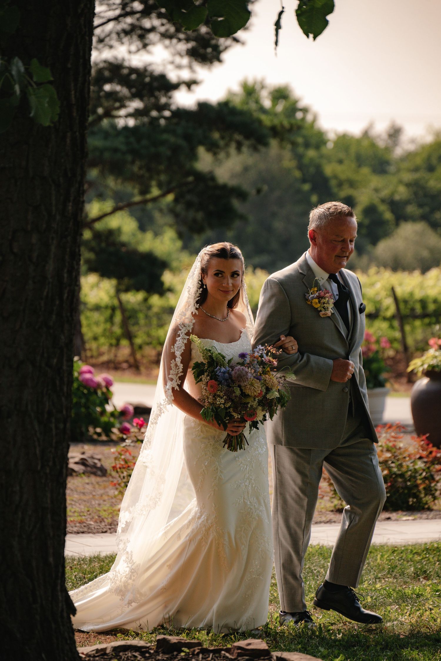 Bride in lace gown holding bouquet walks with suited man through garden vineyard on sunny wedding day.