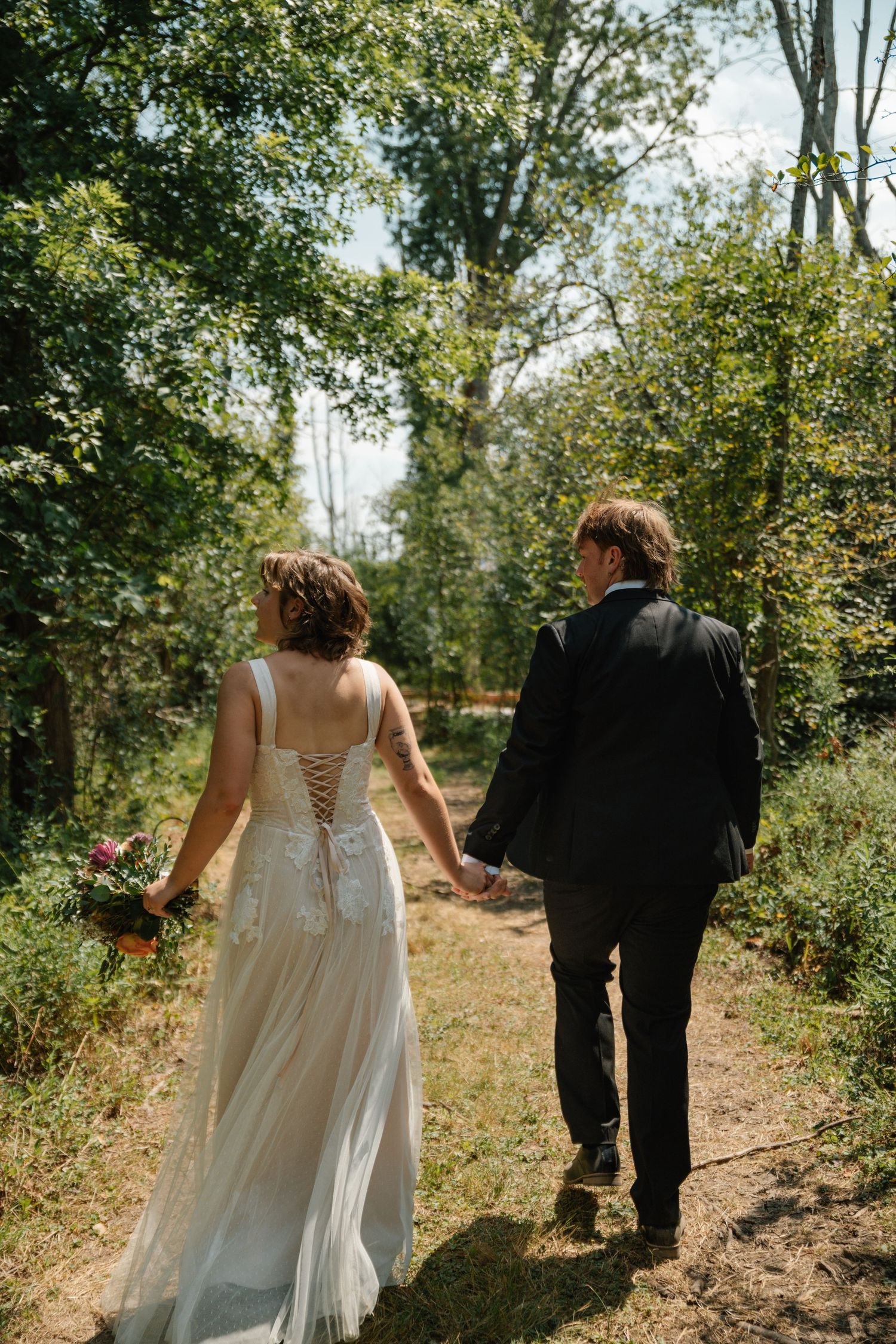 Bride and groom walking hand in hand through a lush green forest path on their wedding day.