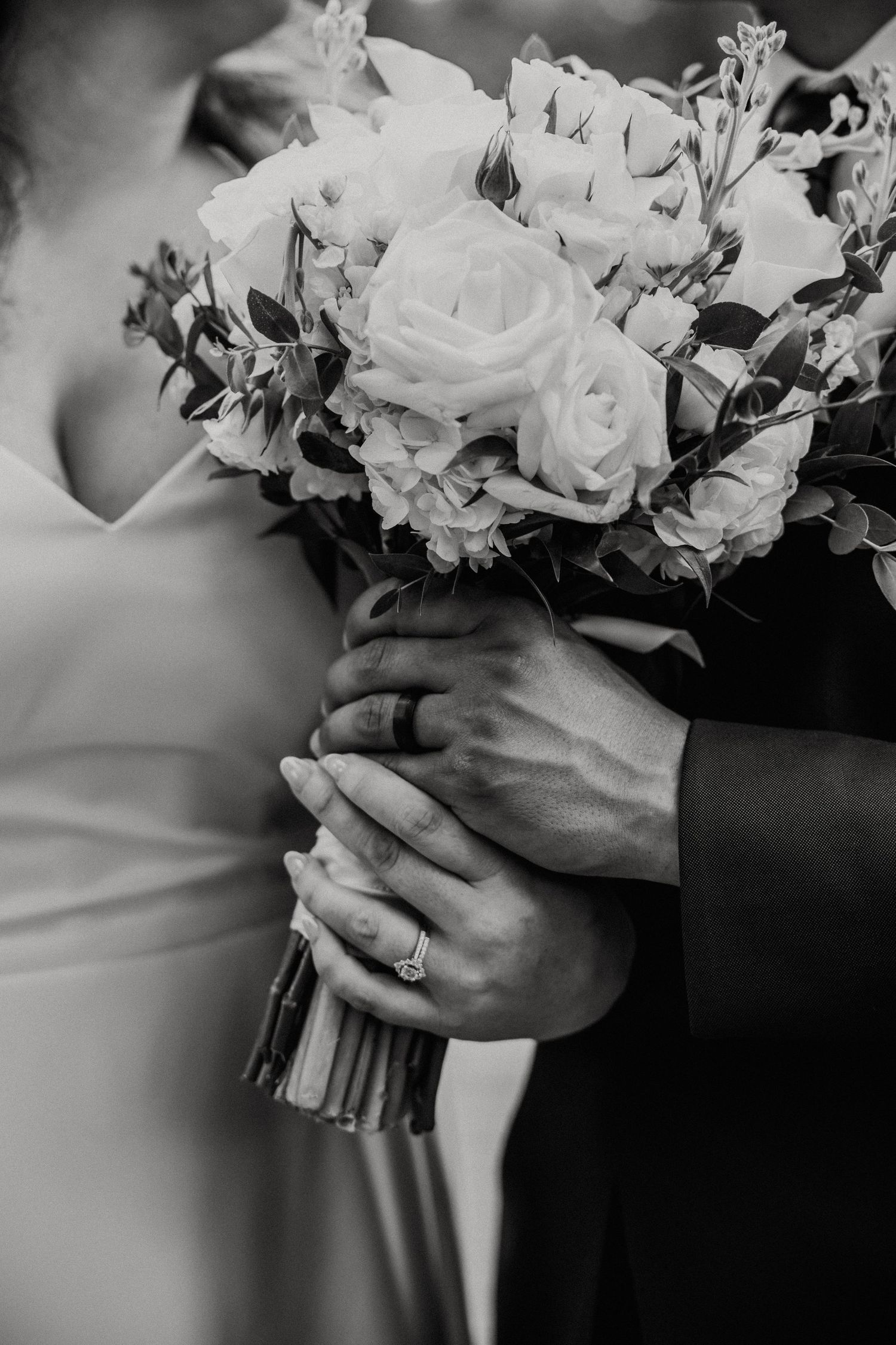 Black and white wedding photo showing hands holding elegant white rose bouquet with an engagement ring visible.