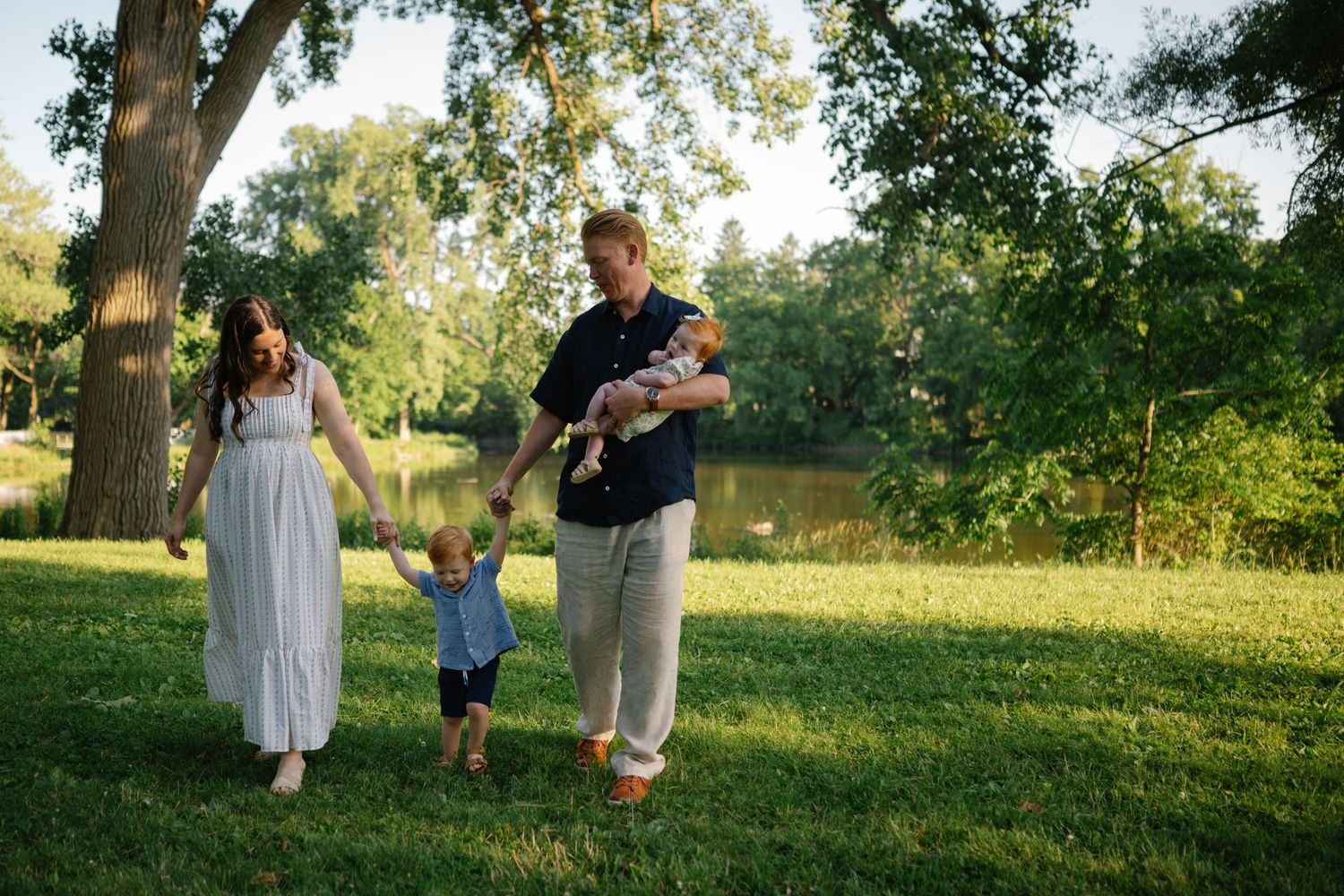Family of four walking outdoors on green grass near trees and a pond on a sunny day.