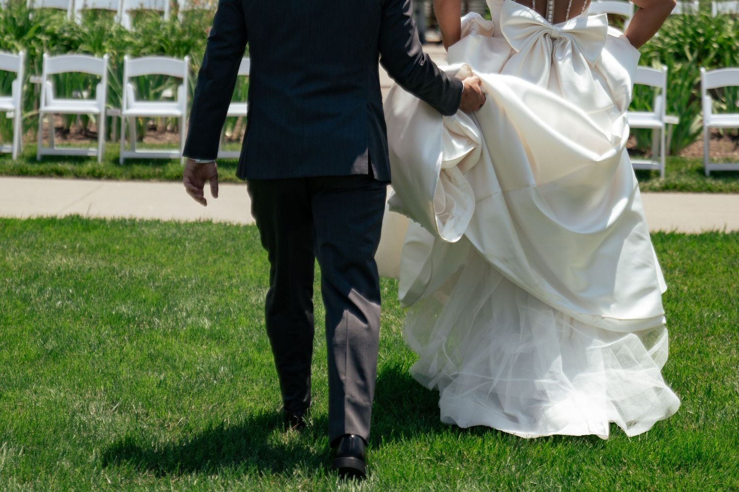 Bride and groom walking on green grass at outdoor wedding ceremony, bride holding white gown train.