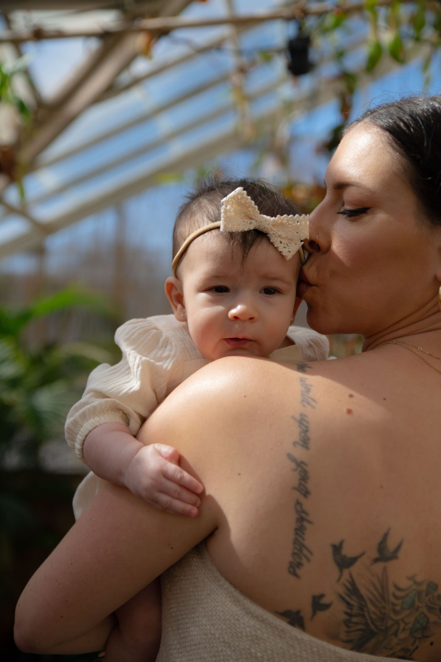 A mother with a tattoo kisses her baby girl wearing a bow headband in a sunlit greenhouse setting.