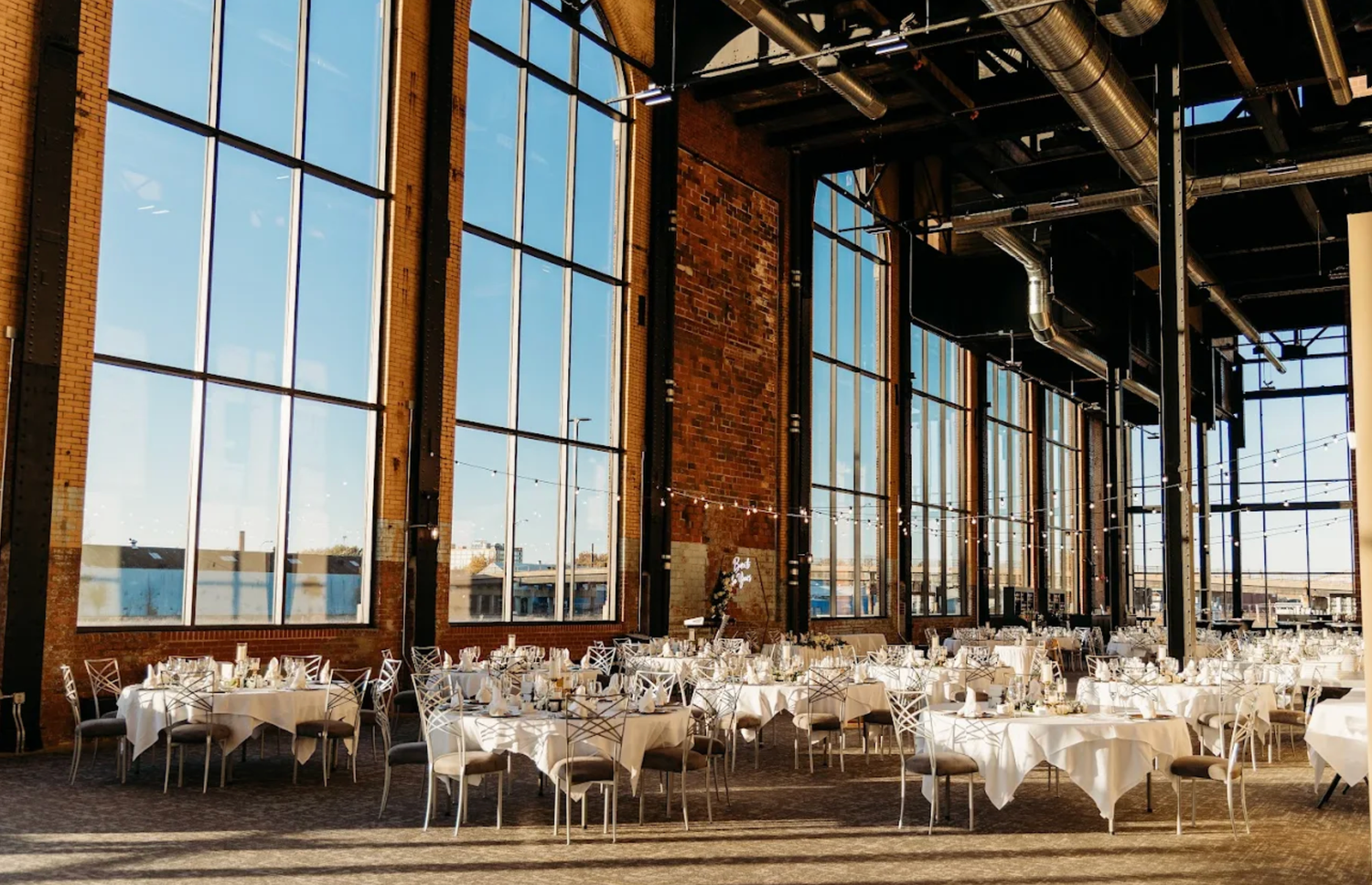 Elegant wedding reception venue with floor-to-ceiling windows, exposed brick walls, and white-clothed dining tables.