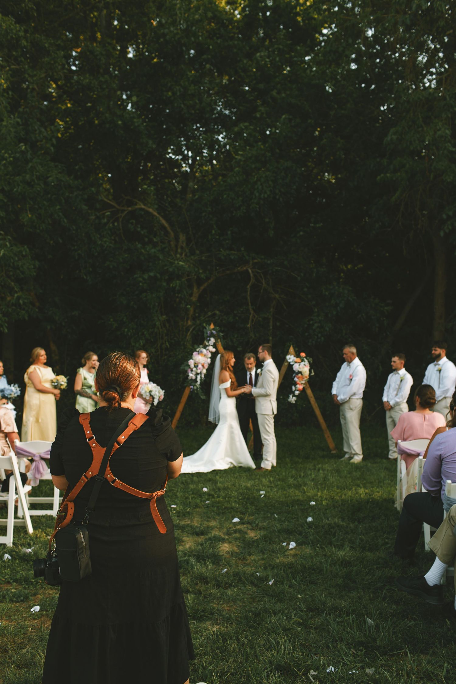 A wedding photographer captures a couple exchanging vows at an outdoor ceremony surrounded by lush green trees.