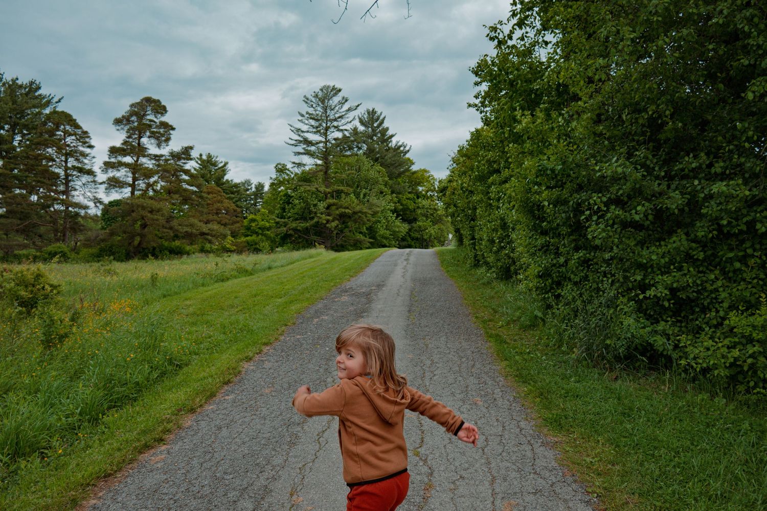A young child in a brown hoodie stands with arms outstretched on a scenic rural gravel road surrounded by trees.