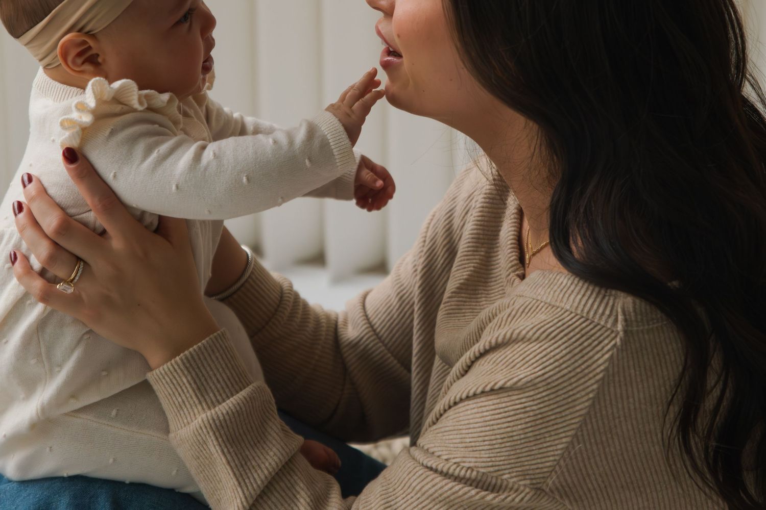 A mother lovingly holds her baby up while the infant touches her face in a warm, tender moment.