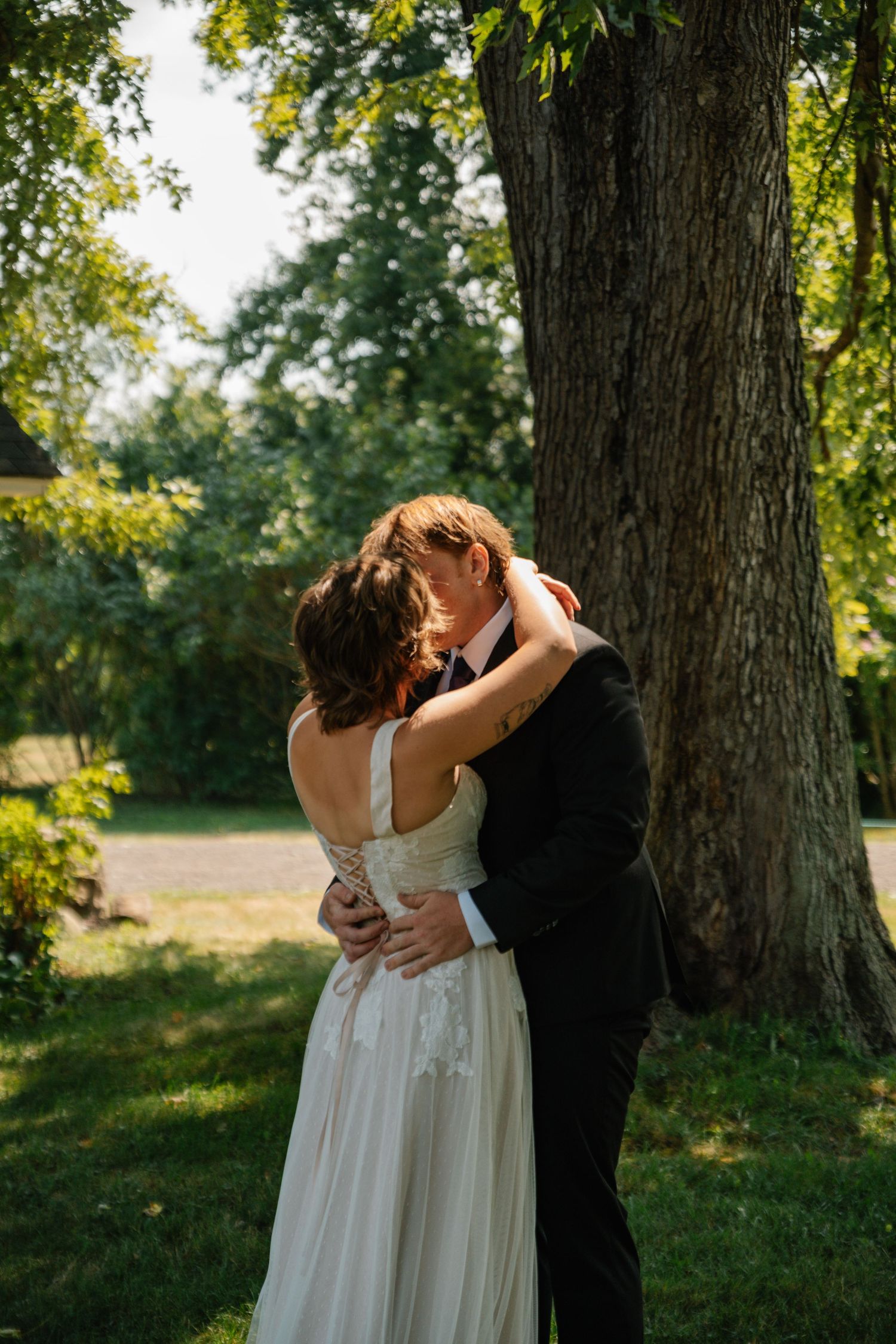 A romantic wedding couple embraces and kisses under a large tree in a sunlit garden setting.