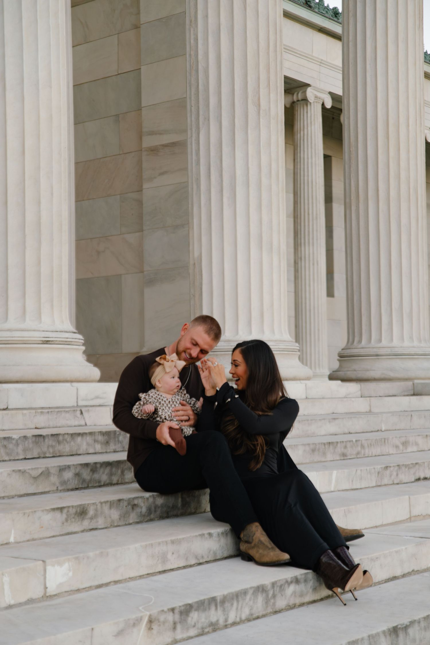 A couple sits on marble steps with columns, lovingly interacting with their baby dressed in a floral outfit.