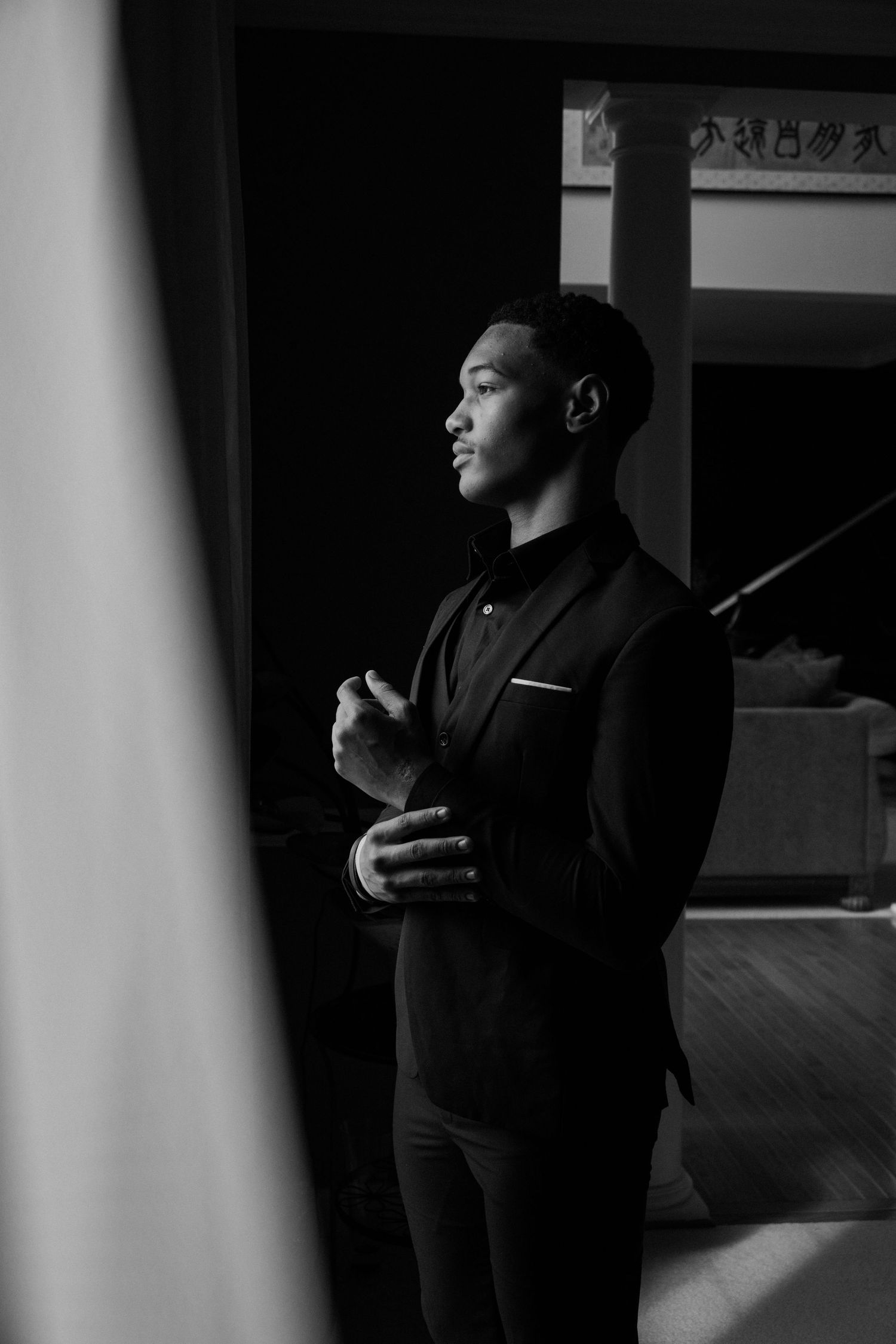 Black and white photo of a young man in a dark suit adjusting his cufflinks, standing in a dimly lit room.