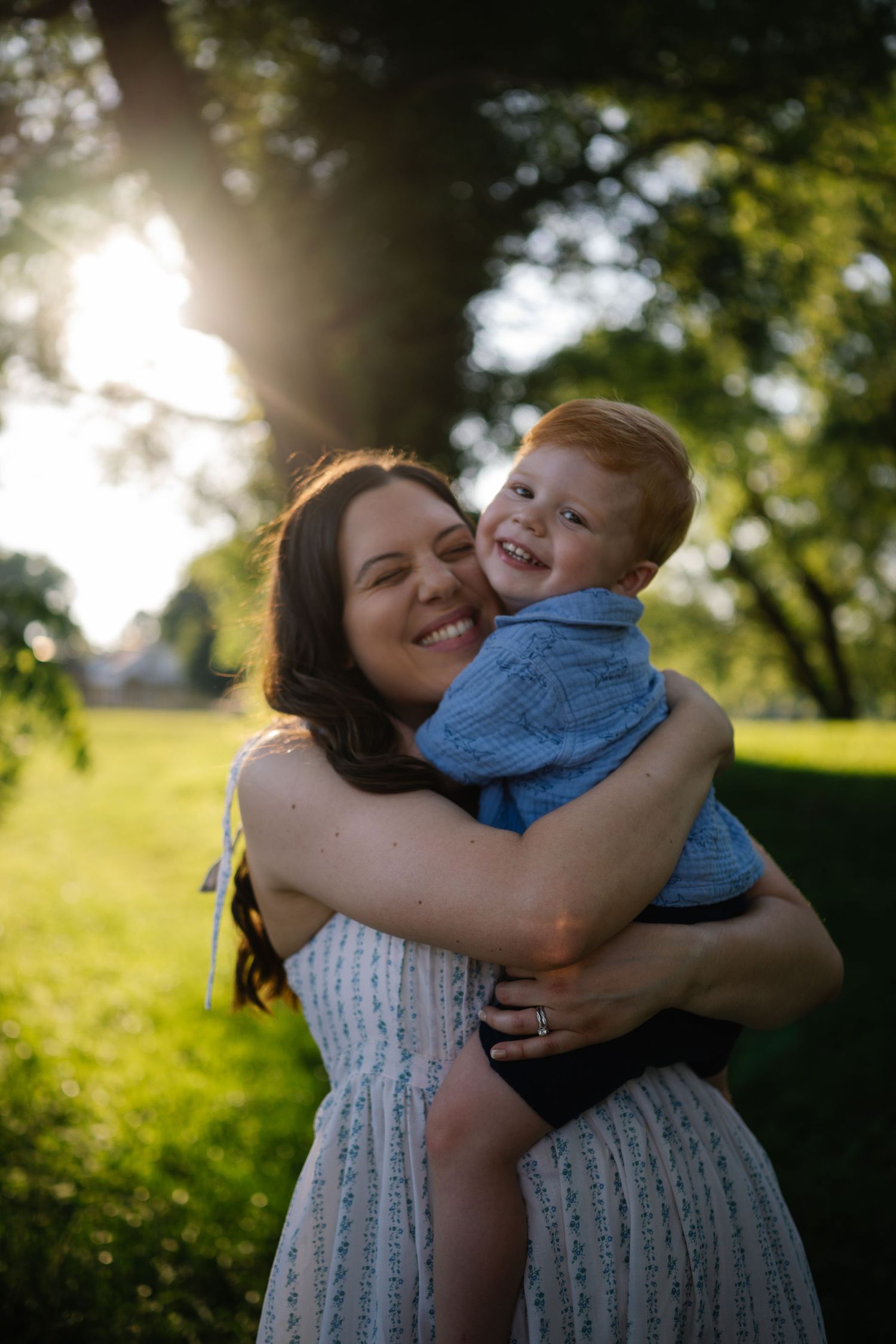 A smiling mother hugs her laughing red-haired boy outdoors in golden sunlight, both radiating joy.