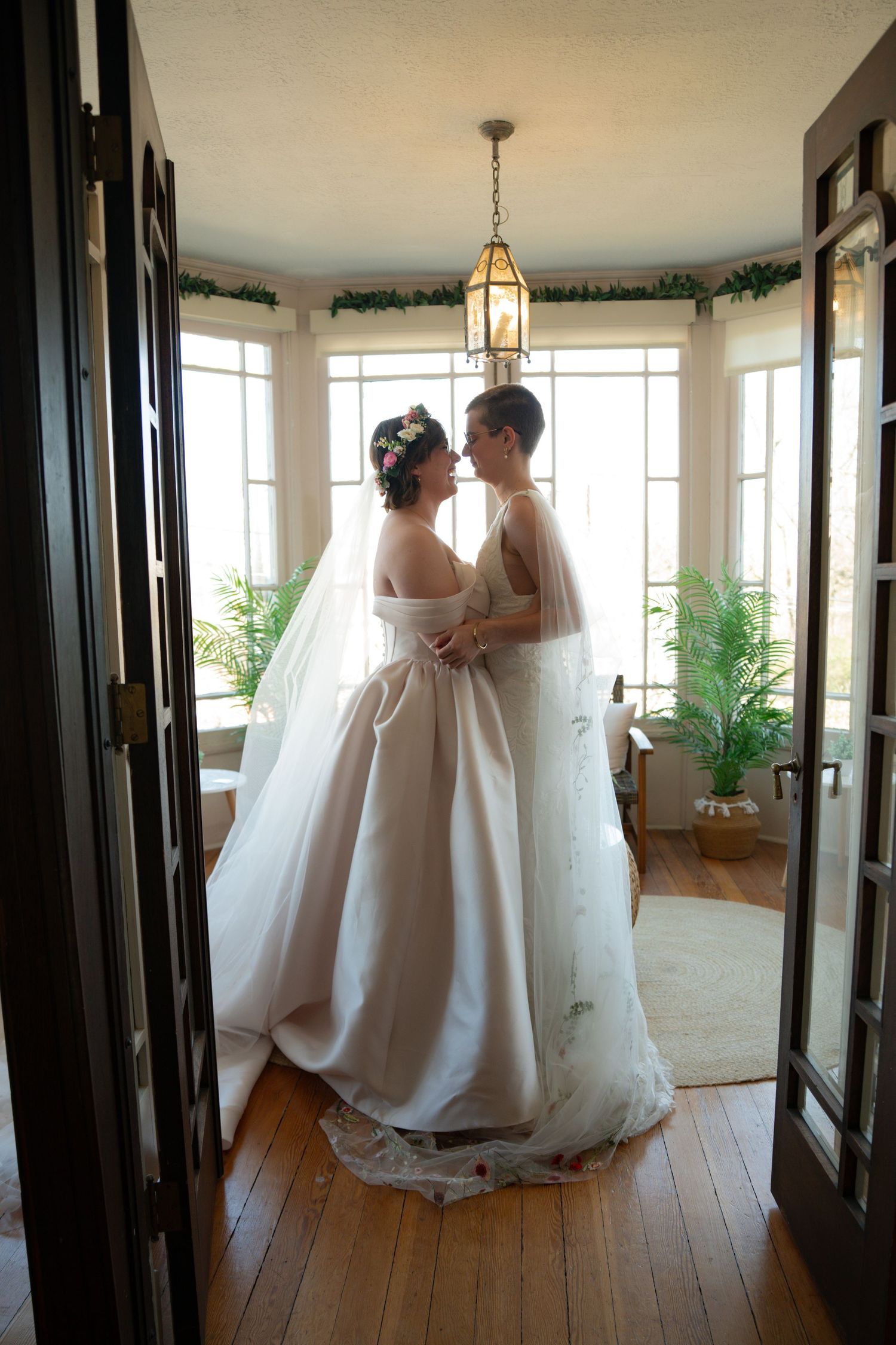 Two brides in white wedding gowns share an intimate moment in a sunlit bay window room with hardwood floors.