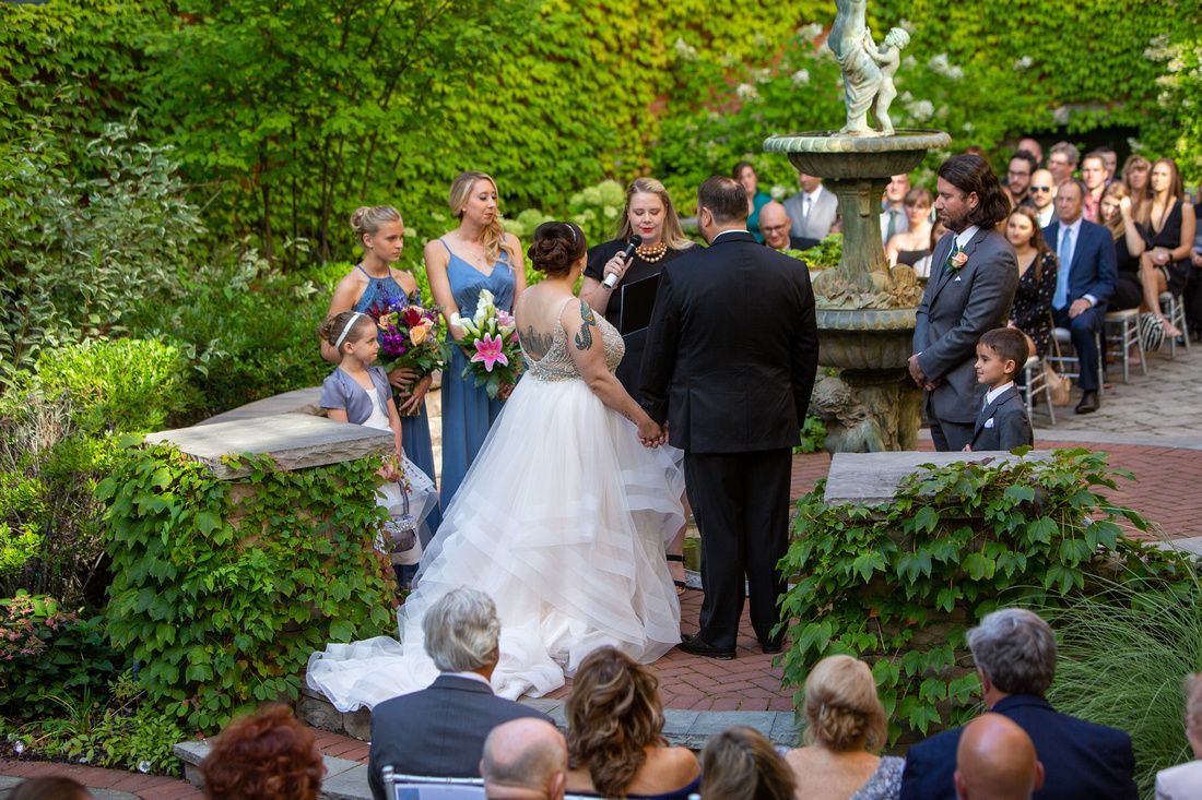 An outdoor garden wedding ceremony takes place near a fountain with guests seated on chairs among lush greenery.