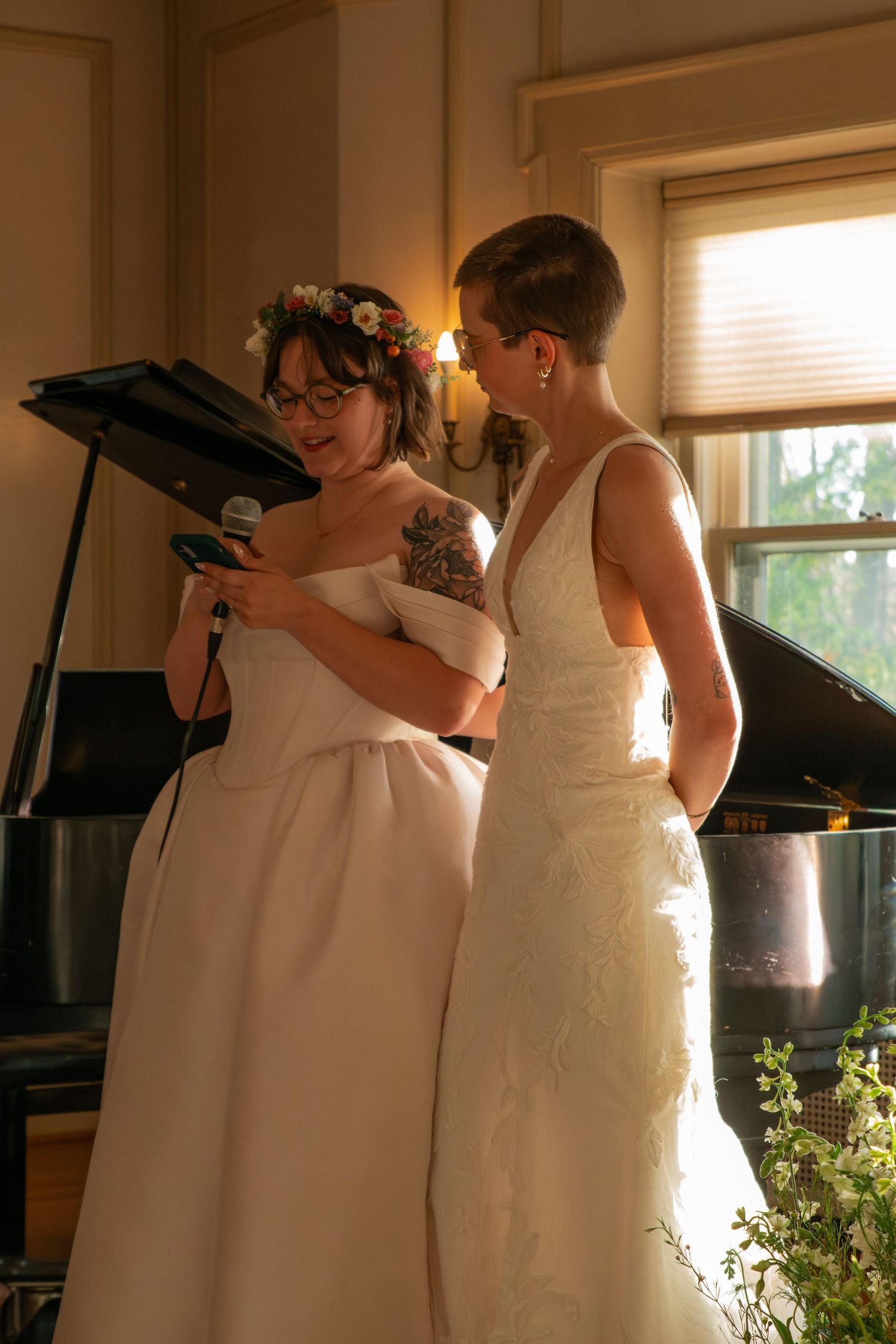 Two brides in white wedding gowns stand together, one holding a microphone near a grand piano.