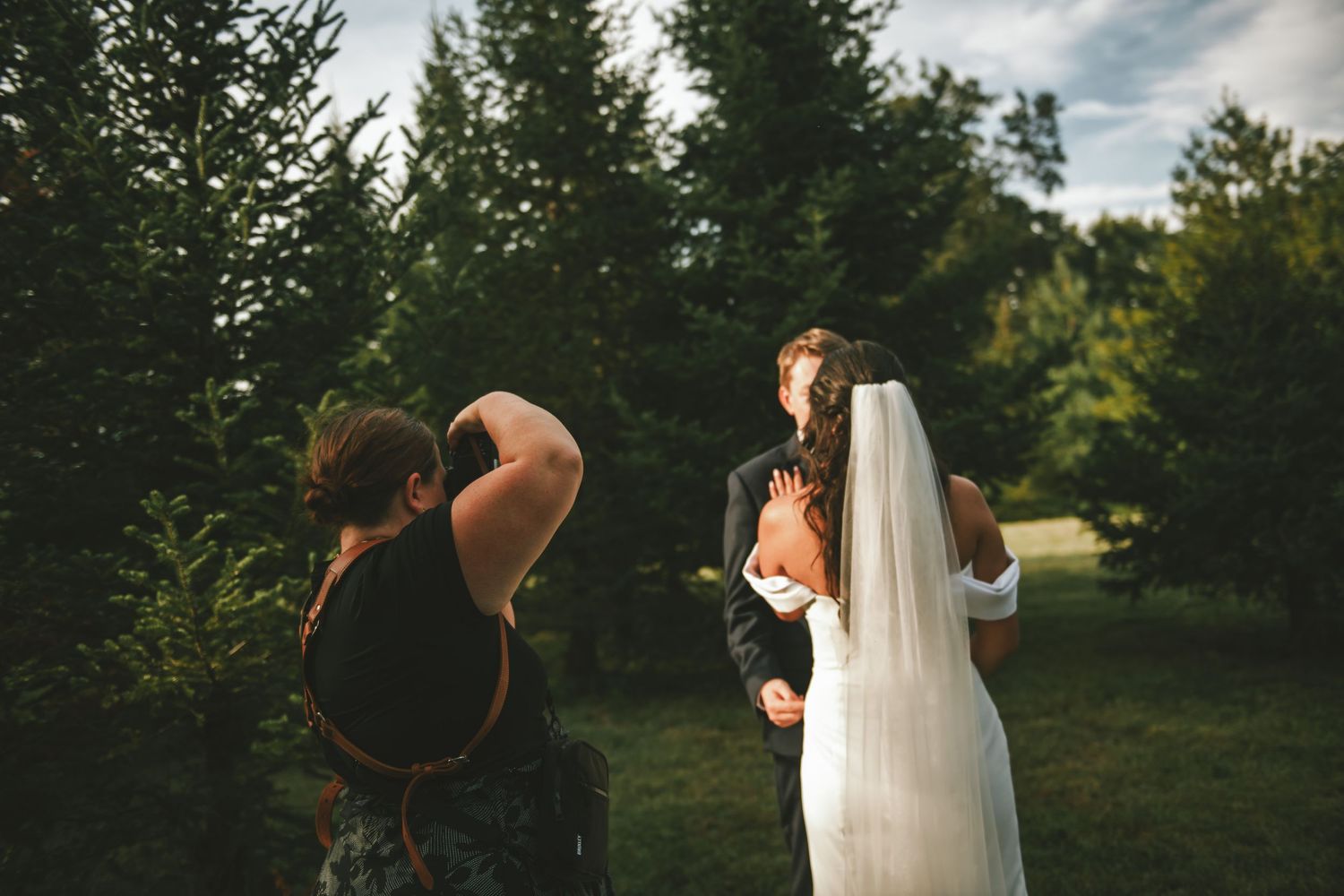 A wedding photographer raises her camera to photograph a bride and groom outdoors among tall evergreen trees.