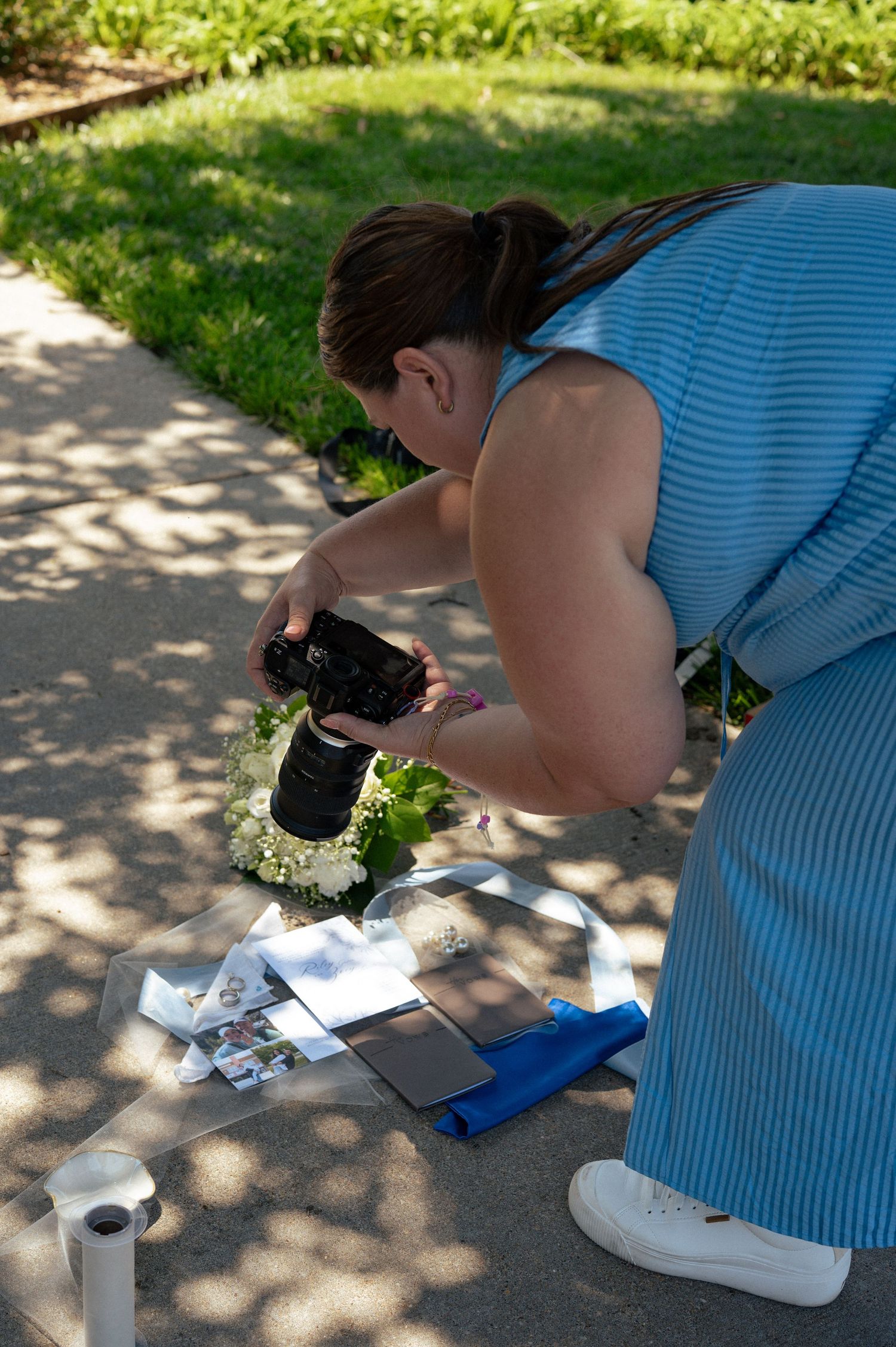 A wedding photographer in a blue dress crouches to photograph floral details and wedding items on a pathway.
