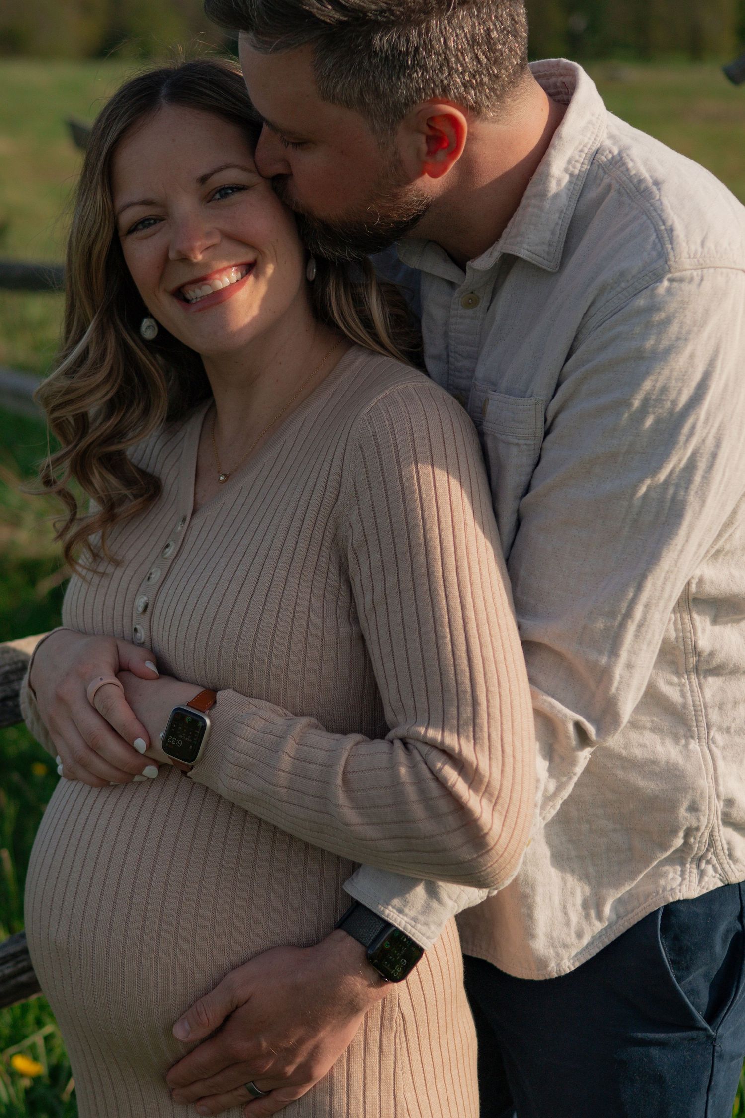 Smiling pregnant woman in beige dress embraced by man kissing her temple outdoors in golden hour light.