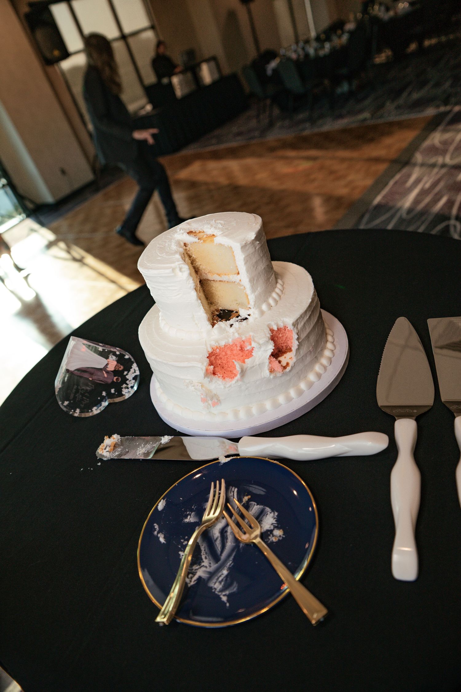 Two-tier white wedding cake with a slice removed, pink flowers, gold forks on navy plate, and cake server on black tablecloth.