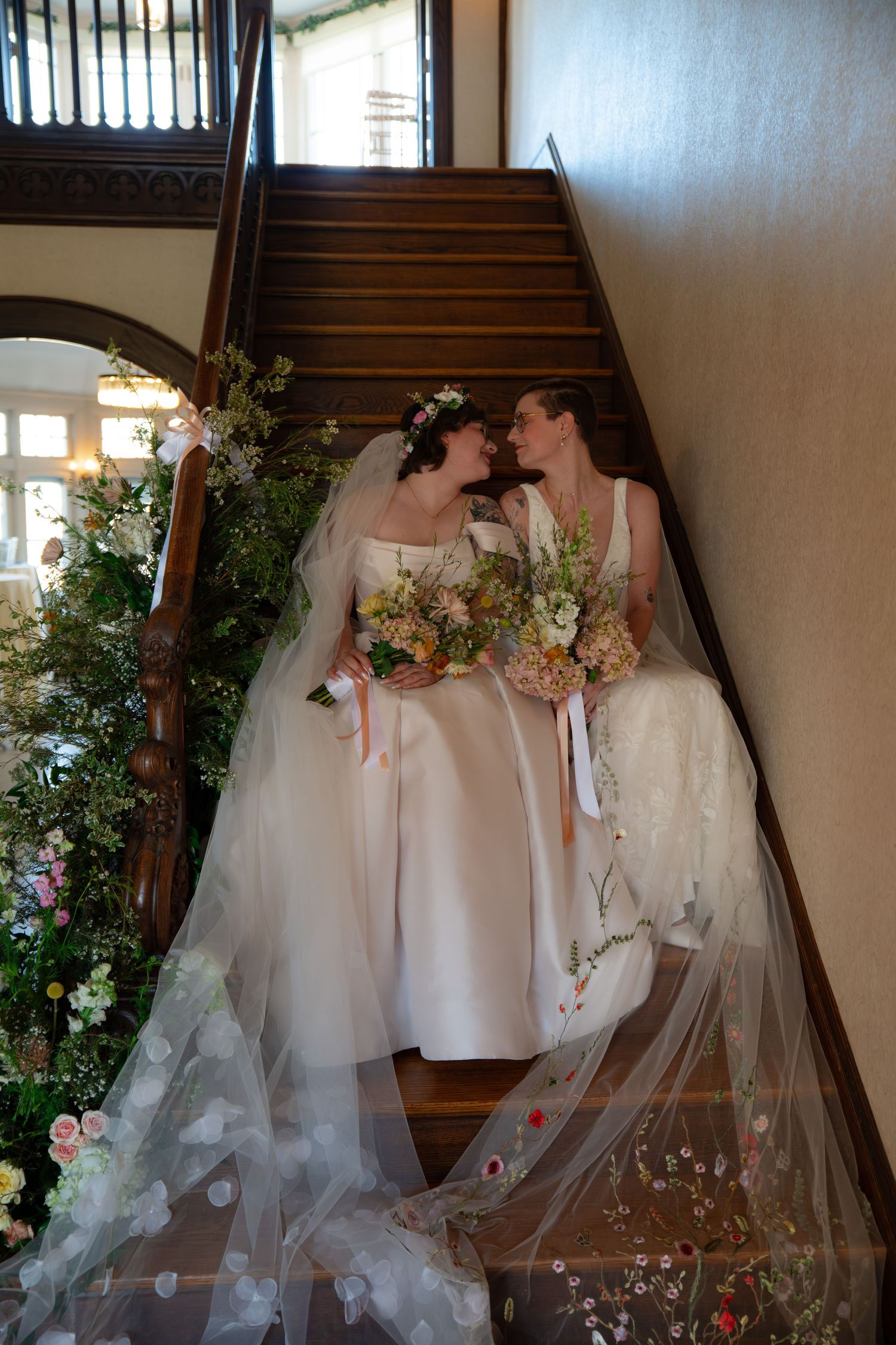Two brides in white gowns with floral bouquets share an intimate moment on a decorated staircase.