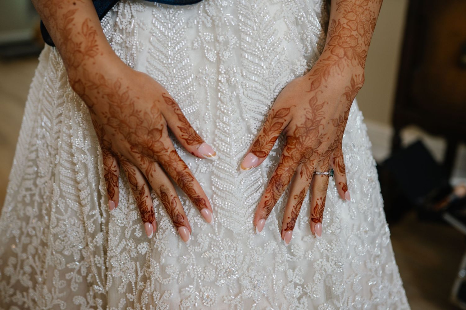 Bride's hands adorned with intricate henna mehndi designs resting on a white lace wedding dress.
