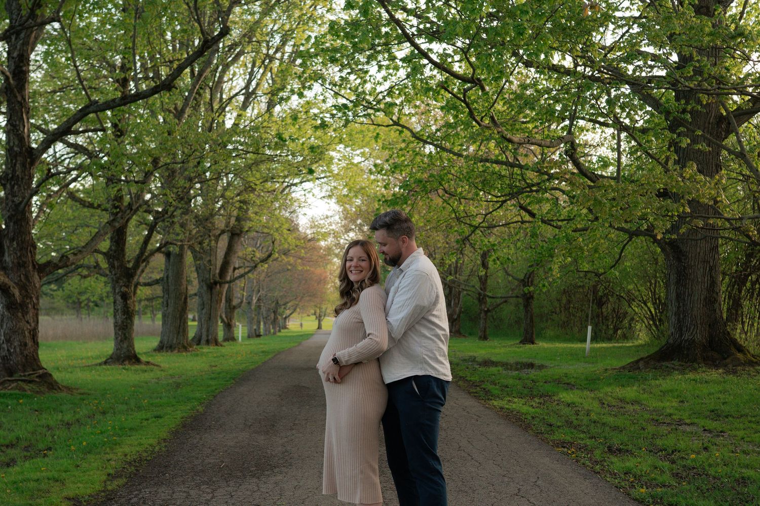 Couple embracing on a tree-lined path during a golden hour maternity photo session outdoors.