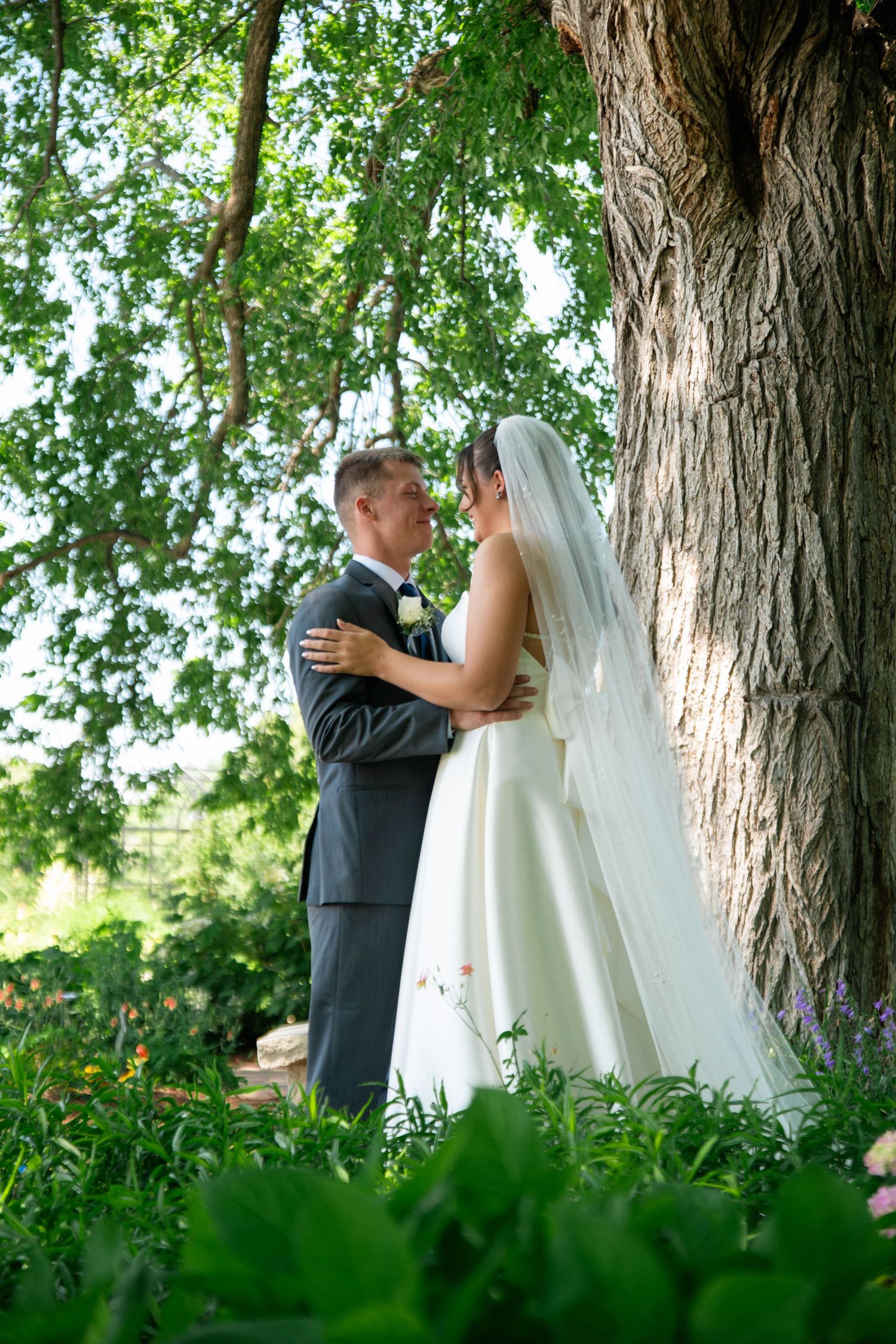 A romantic wedding couple shares an intimate moment under a large tree in a lush green garden setting.