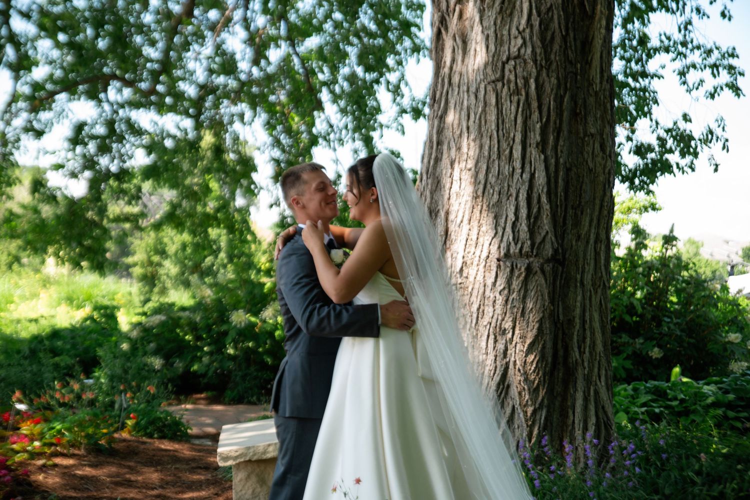 Bride and groom share a romantic kiss beside a large tree in a lush garden on their wedding day.