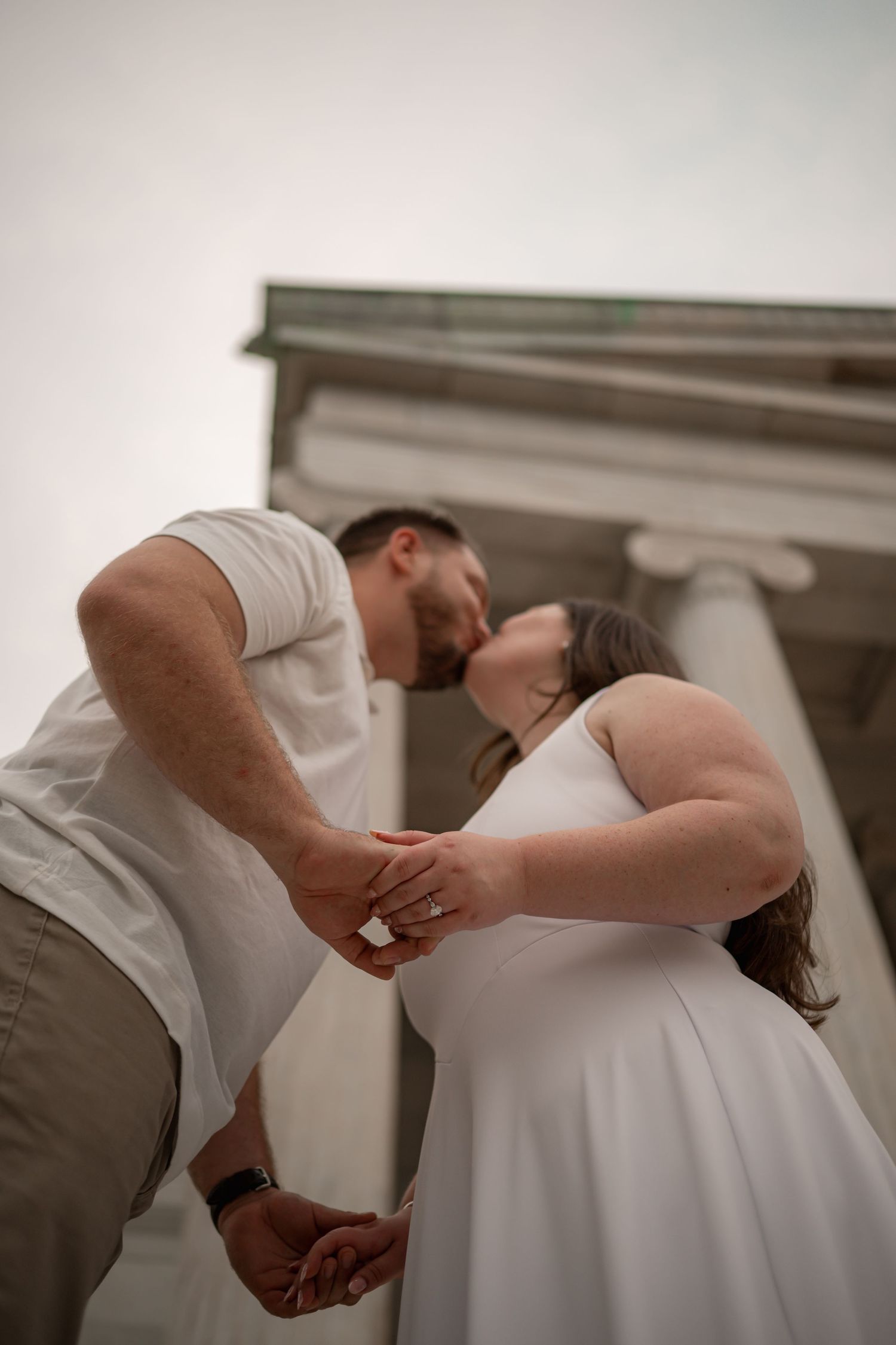 Couple sharing a romantic kiss while holding hands in front of classical columns, woman in white dress, man in white shirt.