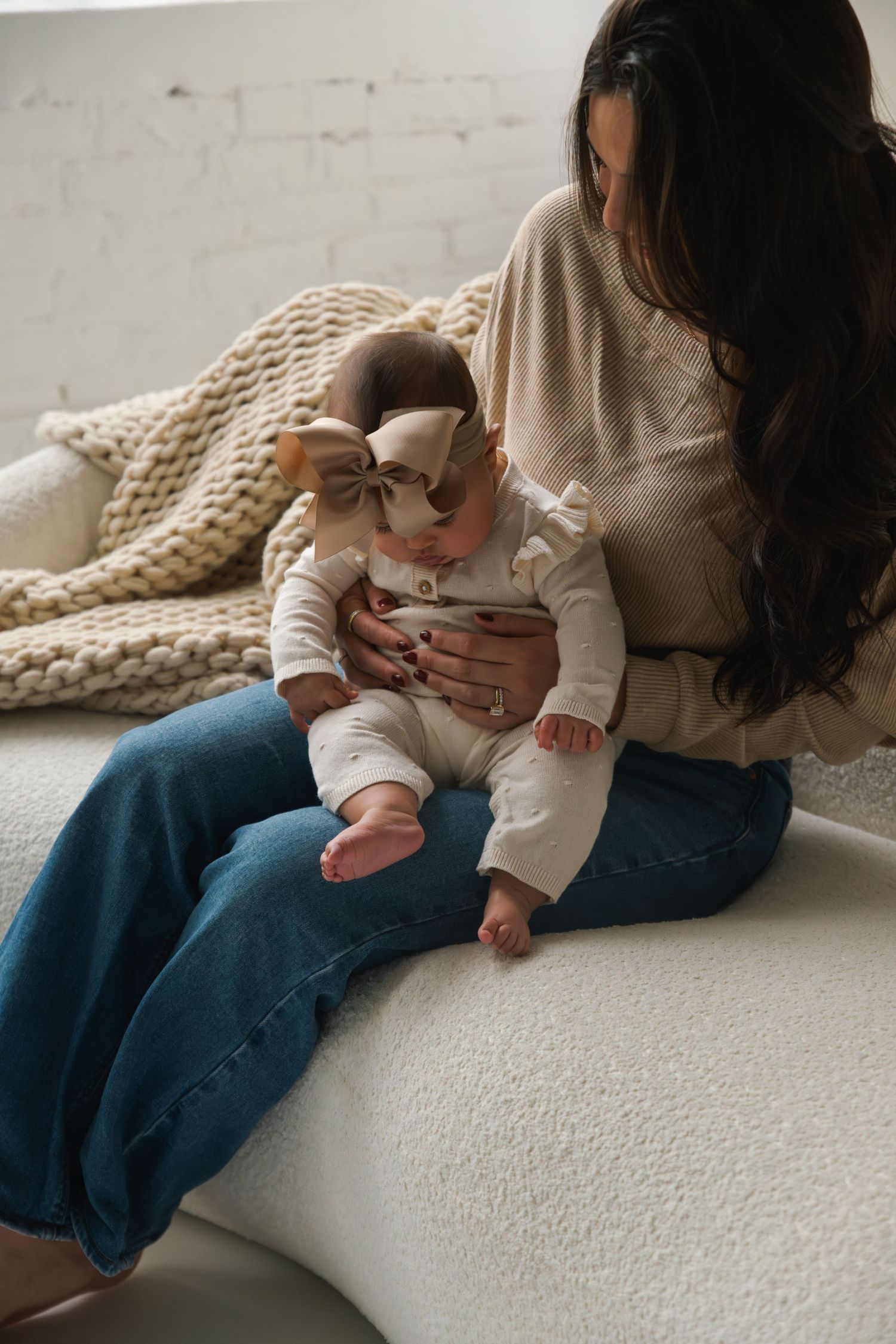A mother in jeans holds her baby girl wearing a bow headband on a white couch with a knit blanket.