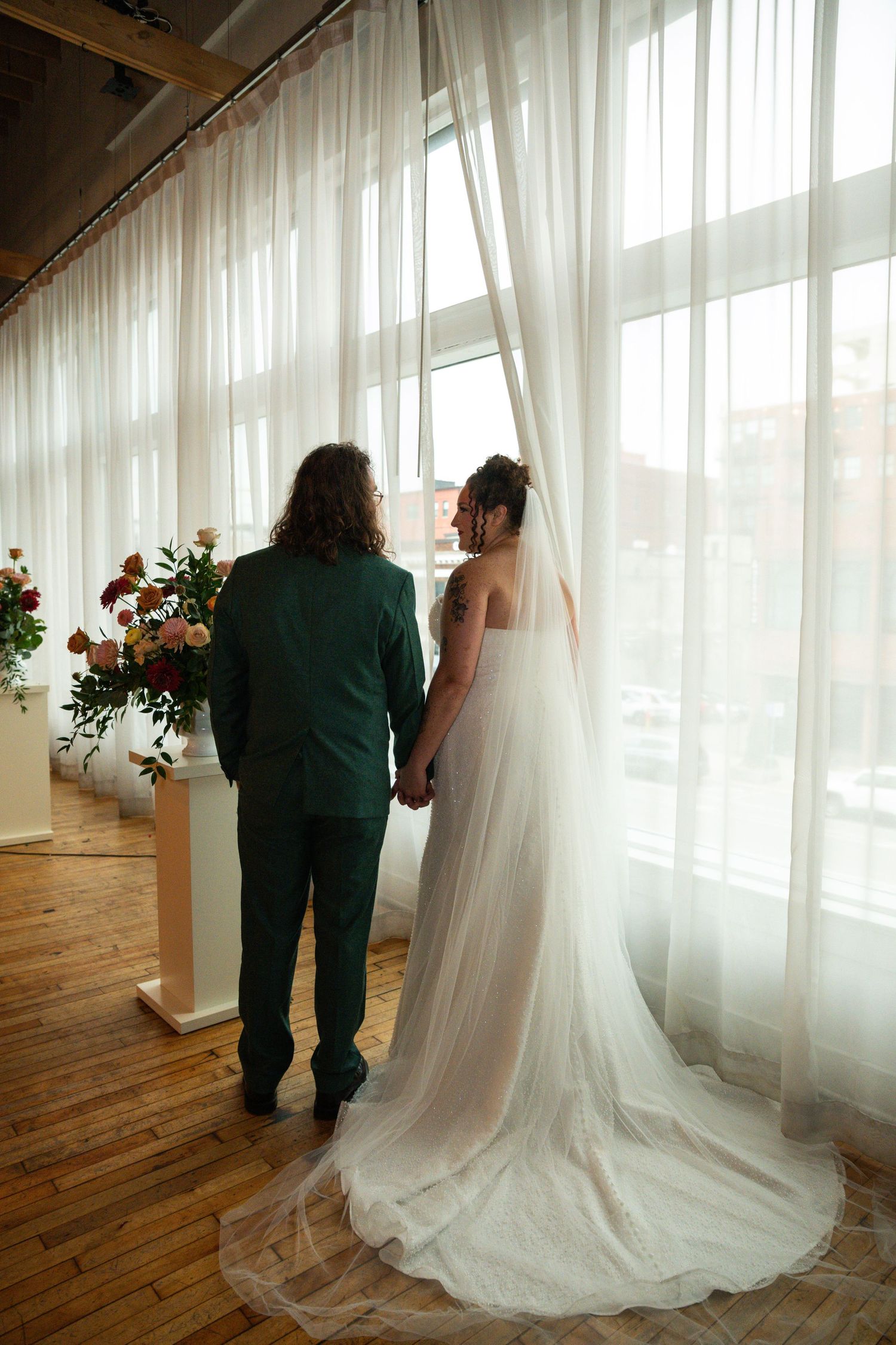 Bride in white gown with veil and groom in green suit hold hands by sheer curtained windows in elegant venue.